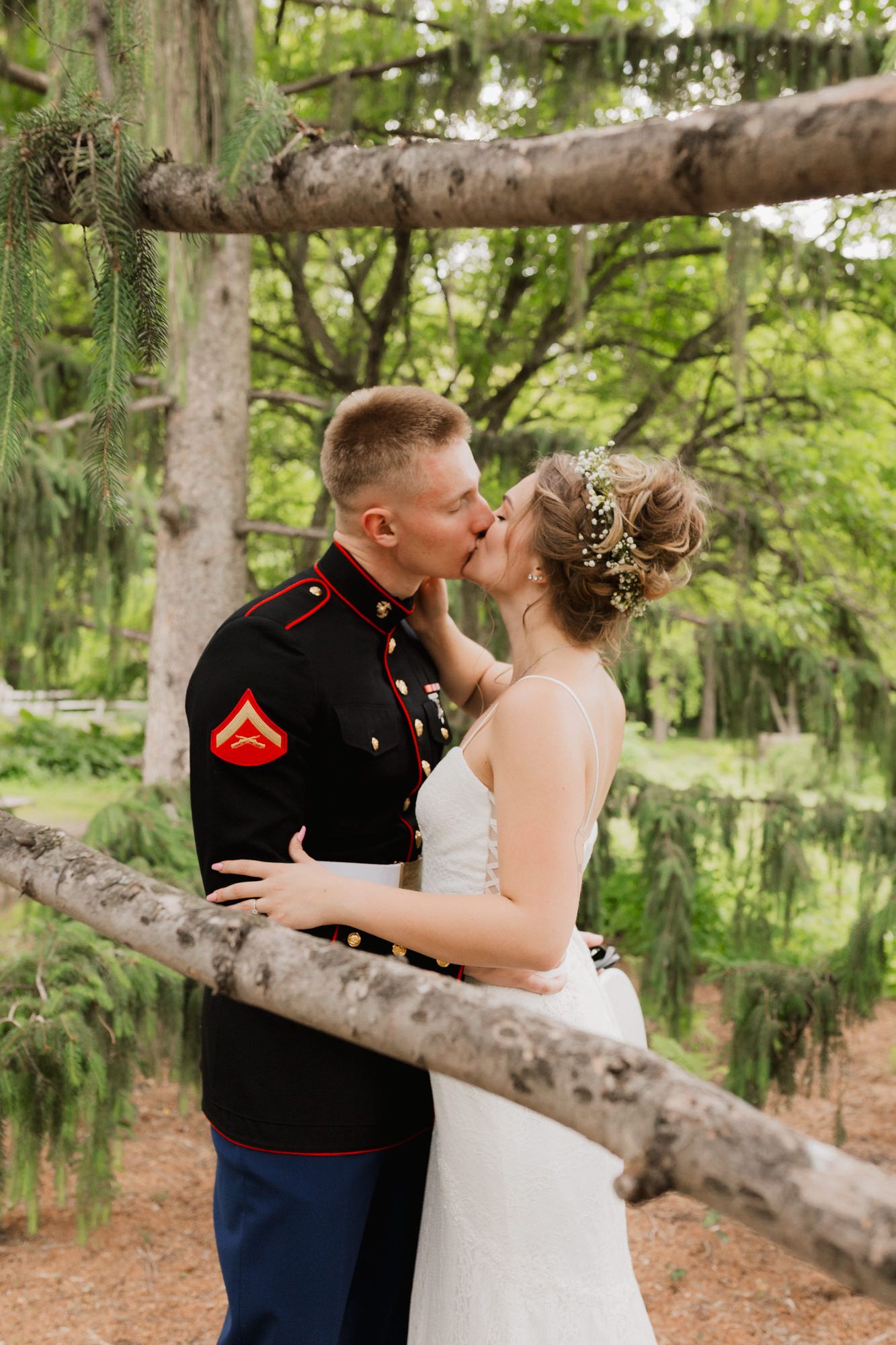 Marine groom in dress blues kisses bride in white gown outdoors among lush green trees at wedding.