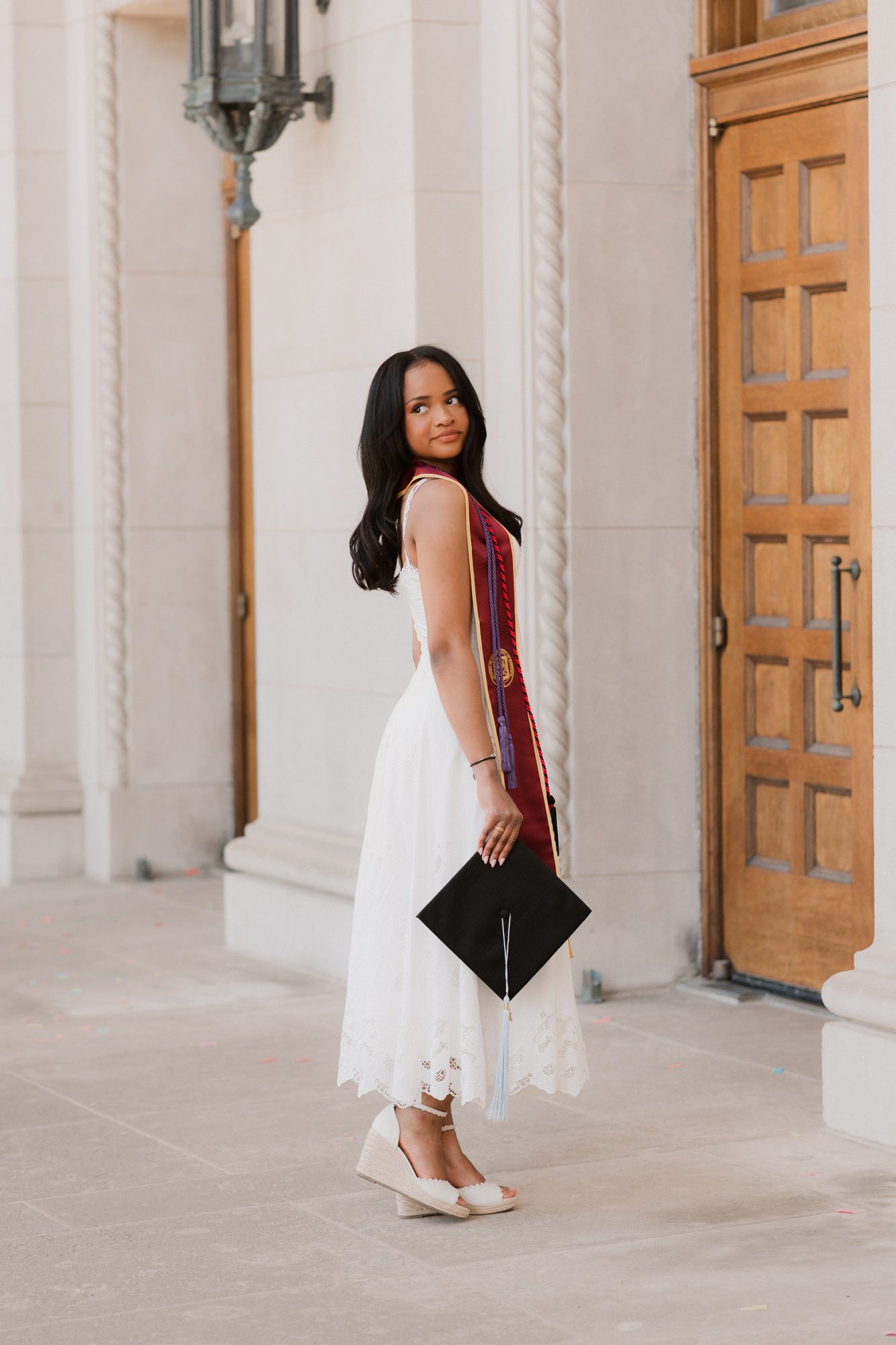 Young woman in white dress and burgundy graduation stole holds cap while posing at elegant building entrance.