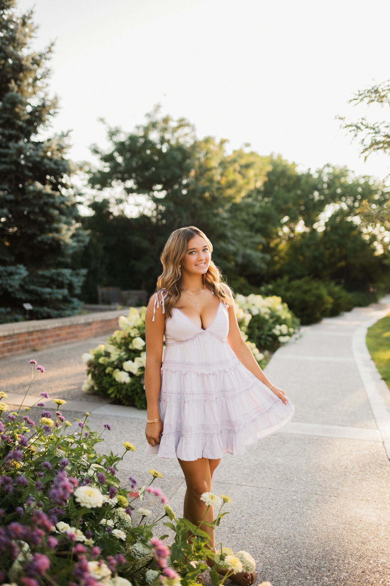 Young woman in white sundress walking along garden path with colorful flowers and green trees in warm sunset light.