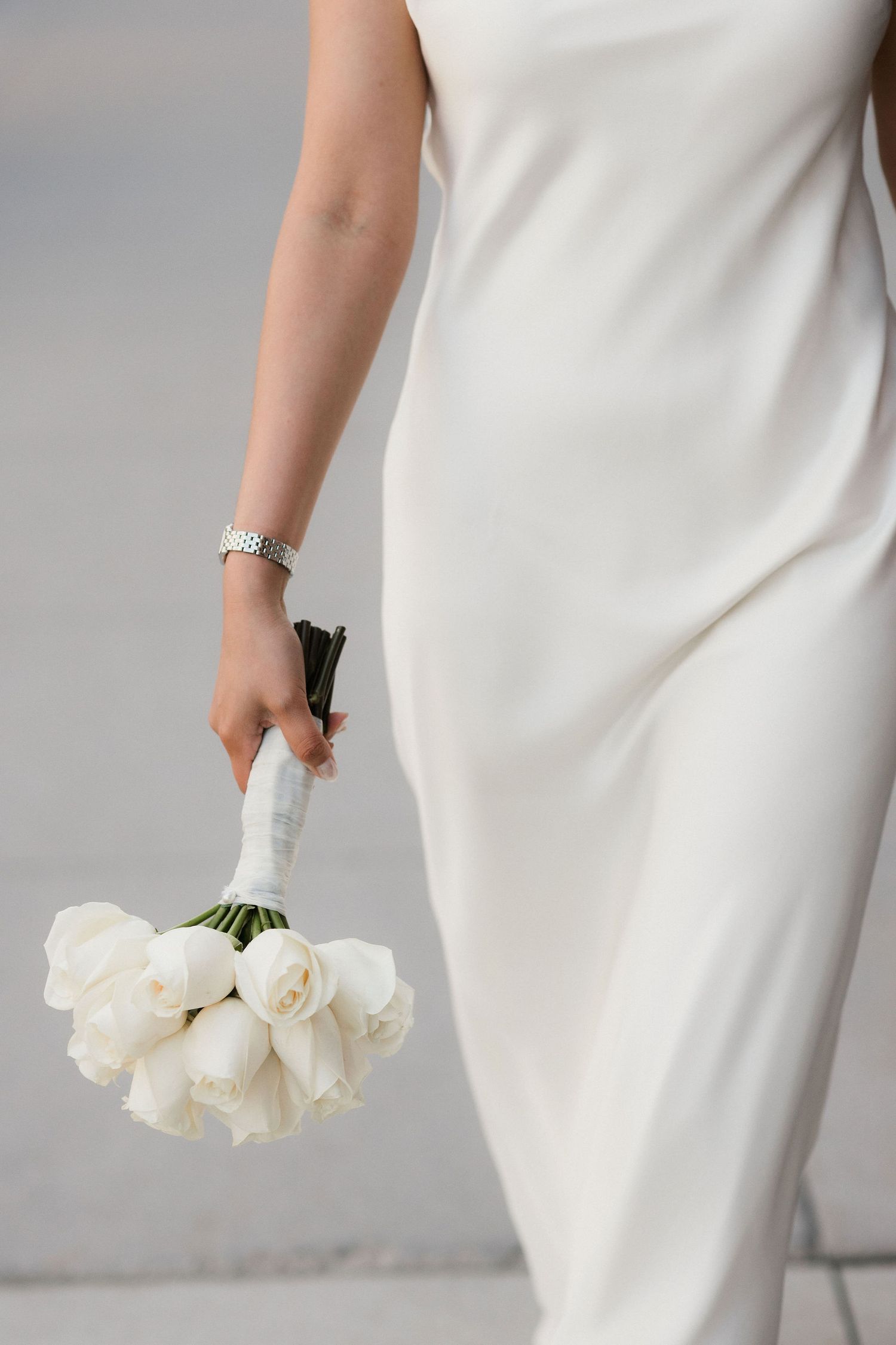 Bride in white satin dress holding elegant bouquet of white tulips tied with ribbon, wearing silver bracelet.