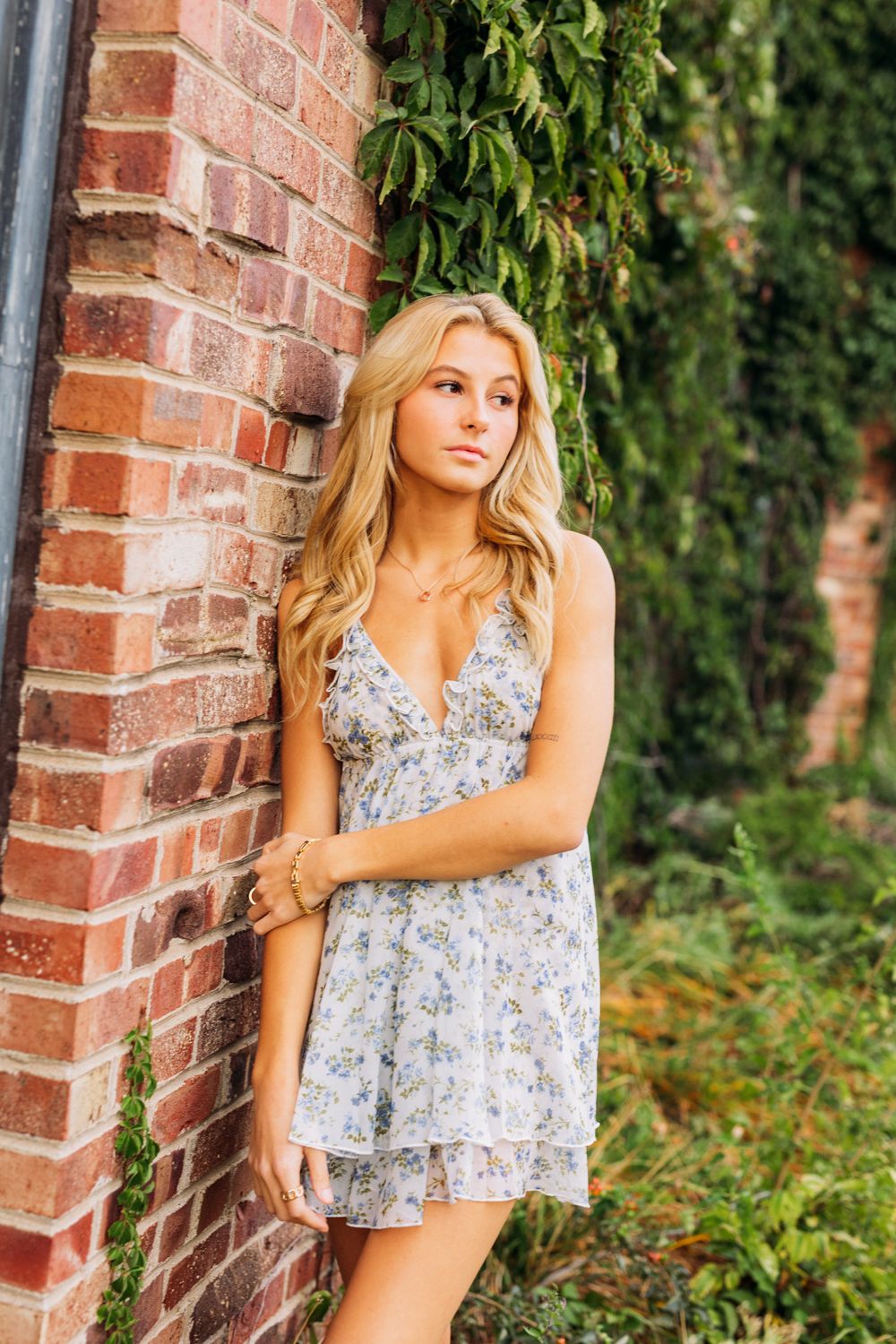 Woman in floral sundress poses against brick wall with ivy climbing up in garden setting.