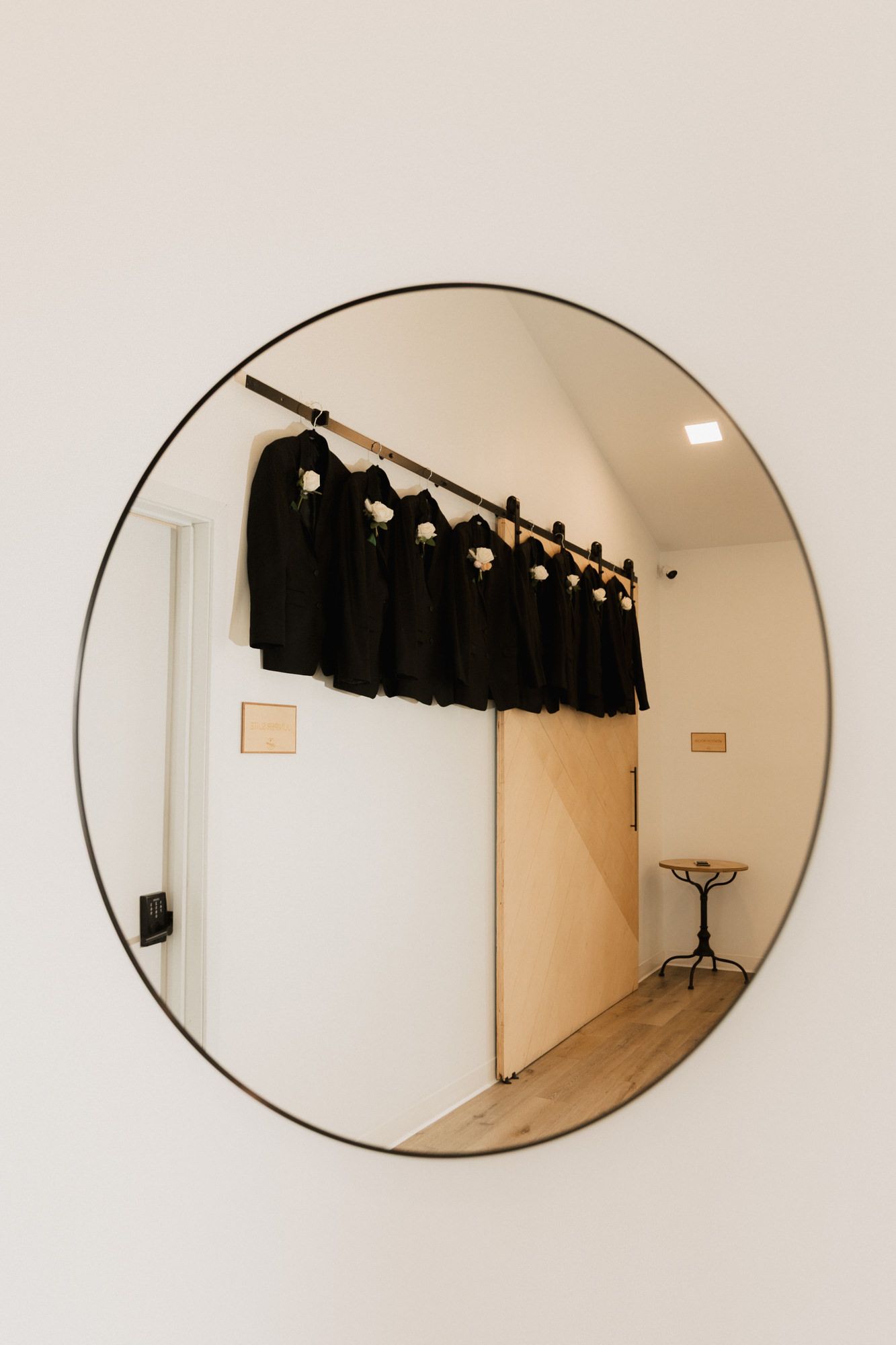 Round mirror reflecting black suits with white flowers on hangers in a bright wedding preparation room.