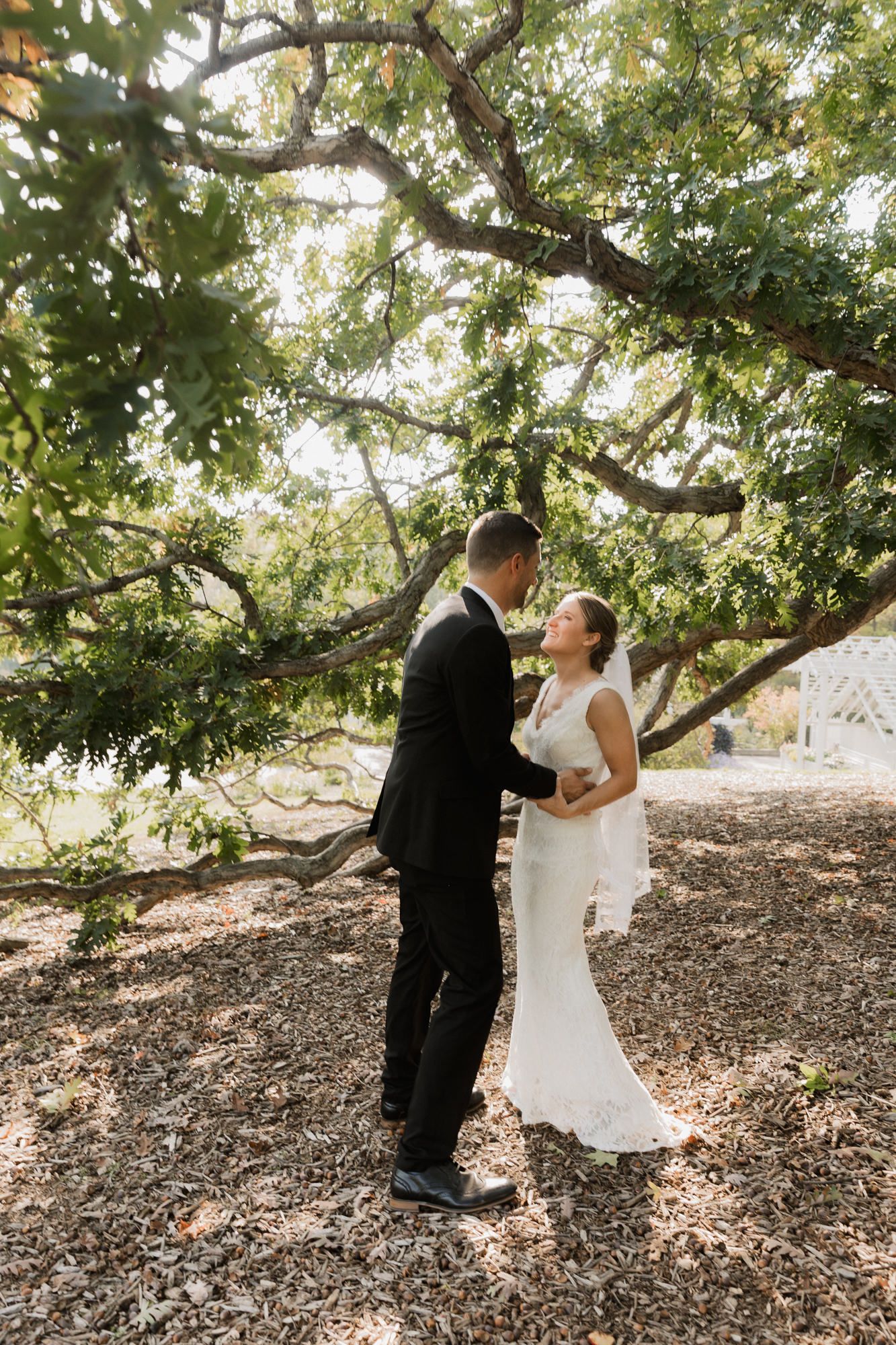 A bride and groom share a romantic moment under a large tree, with the bride in a white gown and groom in a black suit.
