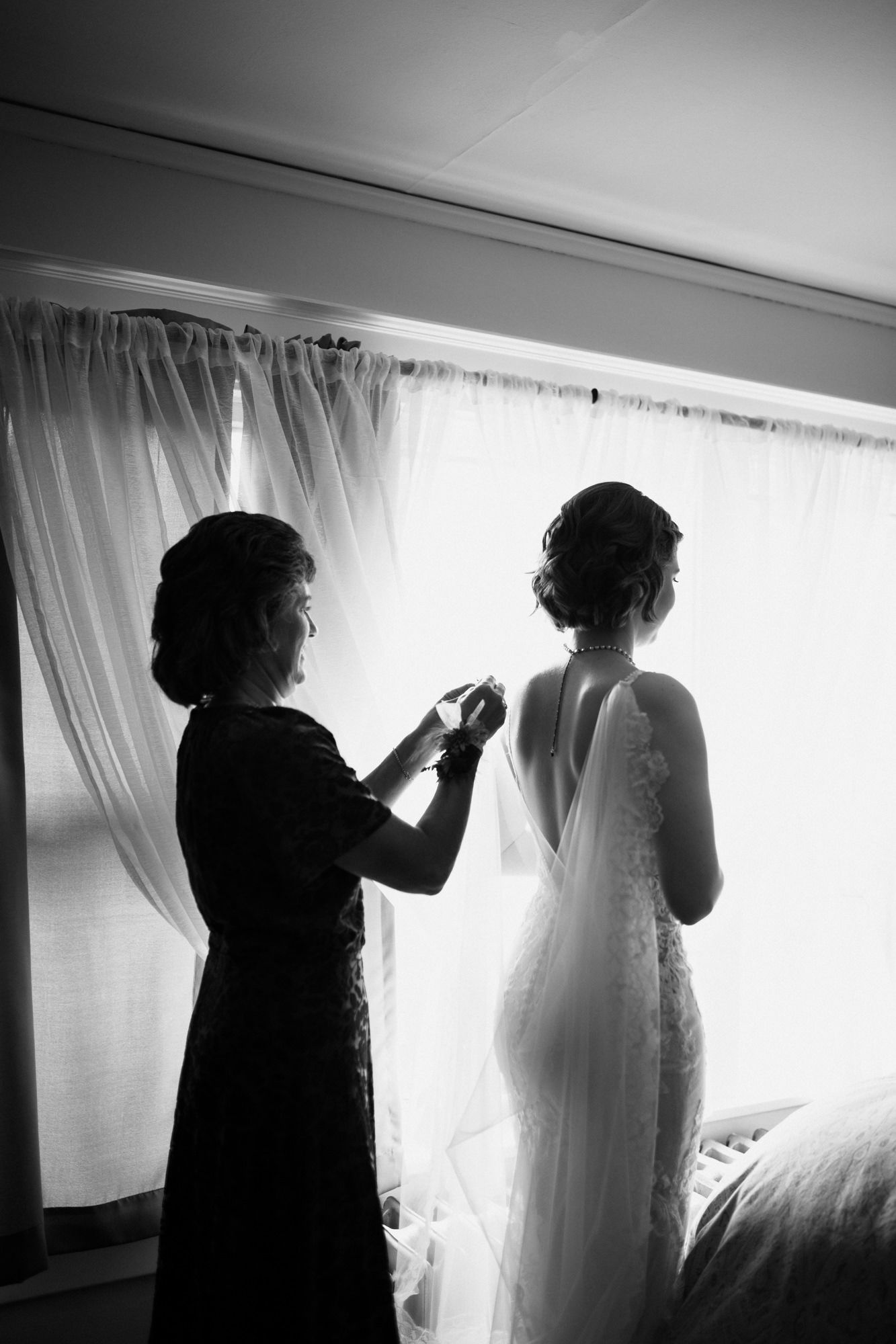 Black and white photo of mother helping bride button wedding dress by window.