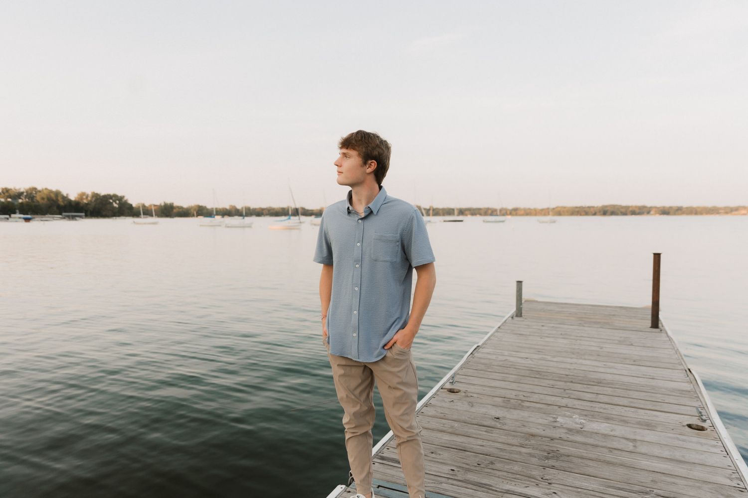 Young man in blue button-up shirt and khaki pants standing on wooden dock overlooking calm lake.