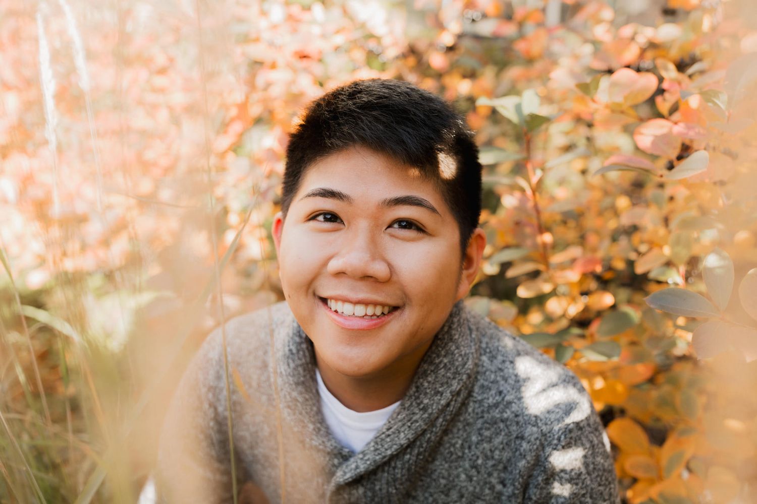 Smiling young person with short dark hair surrounded by autumn foliage in warm golden tones.