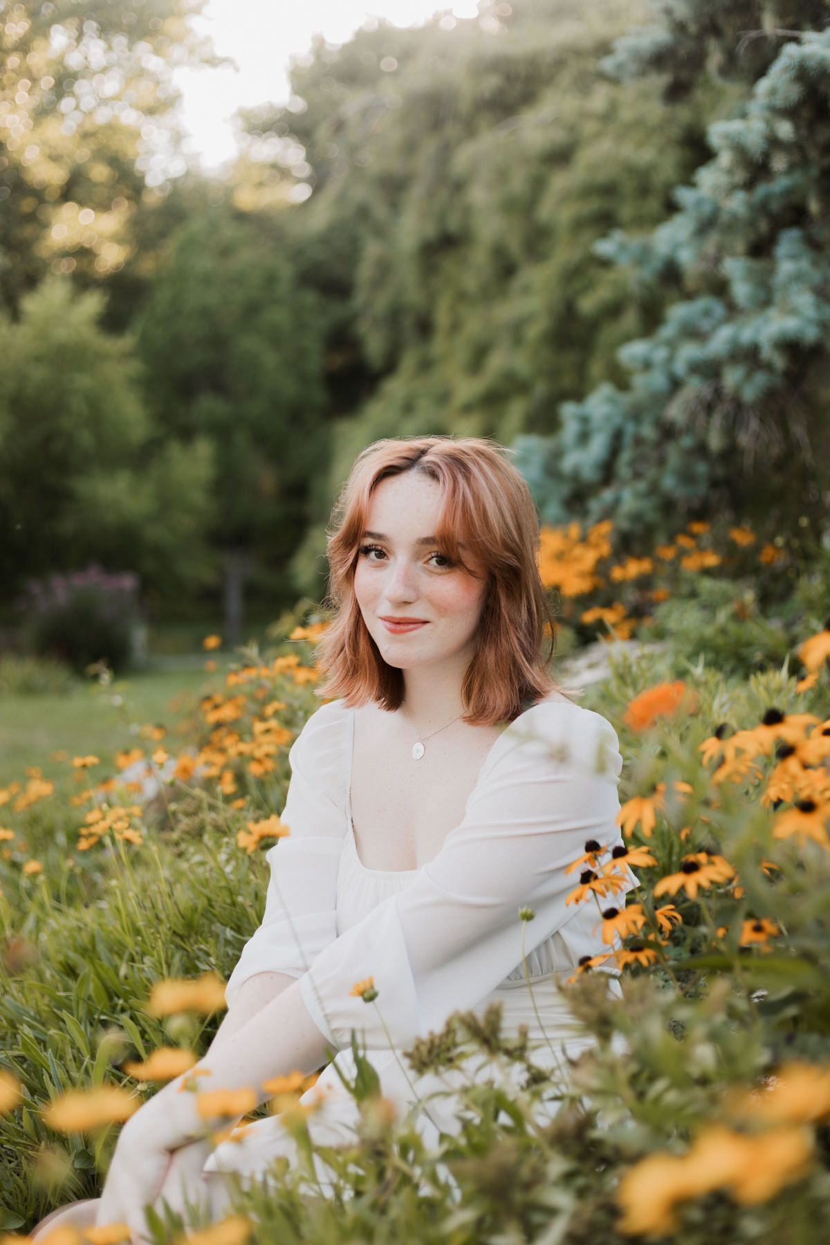 Young woman with auburn hair in white dress sitting among yellow wildflowers in a lush green garden.