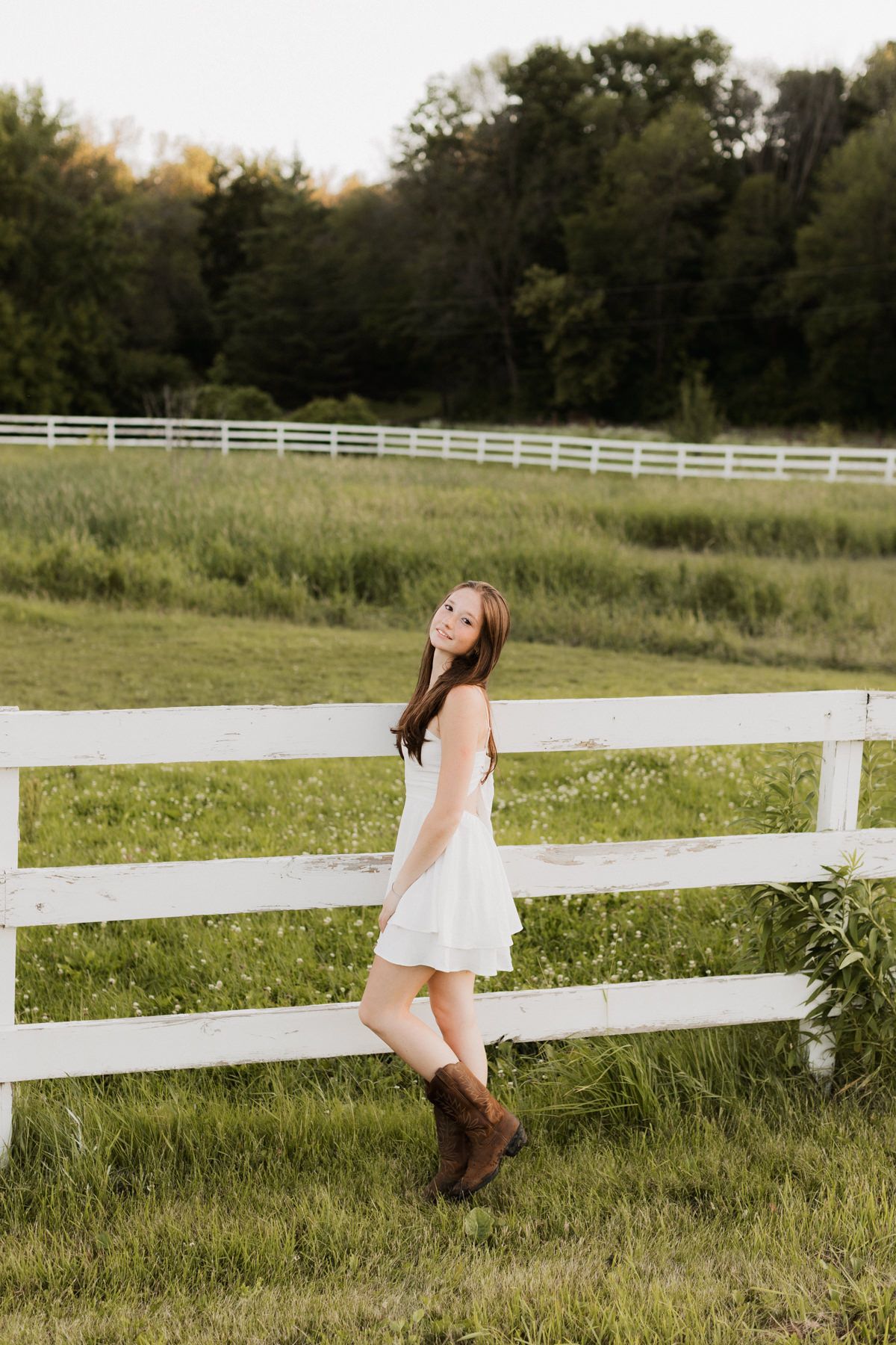 Young woman in white dress and cowboy boots leaning on white farm fence in green field.