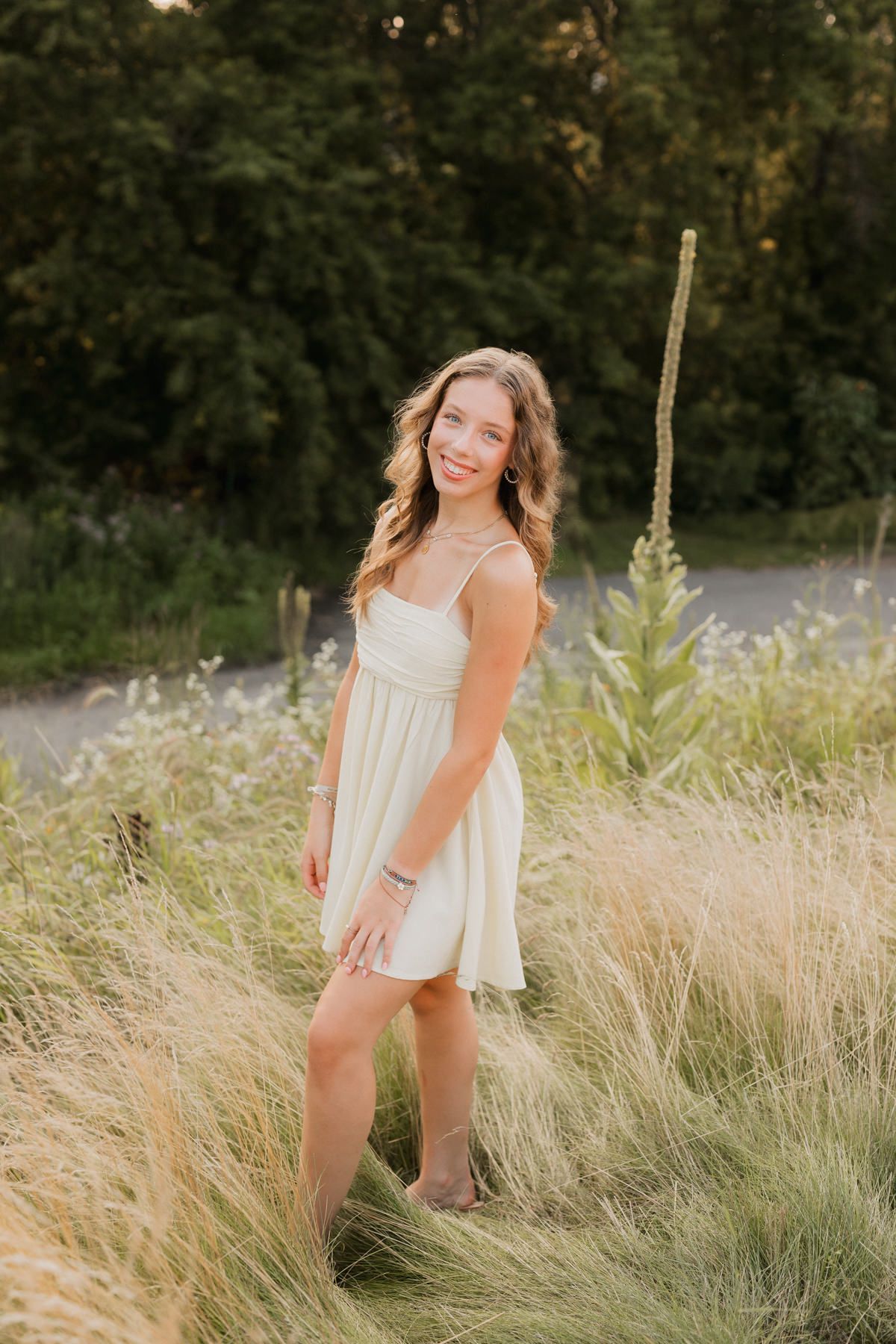 Young woman in white sundress standing in tall grass with green trees in background during golden hour.