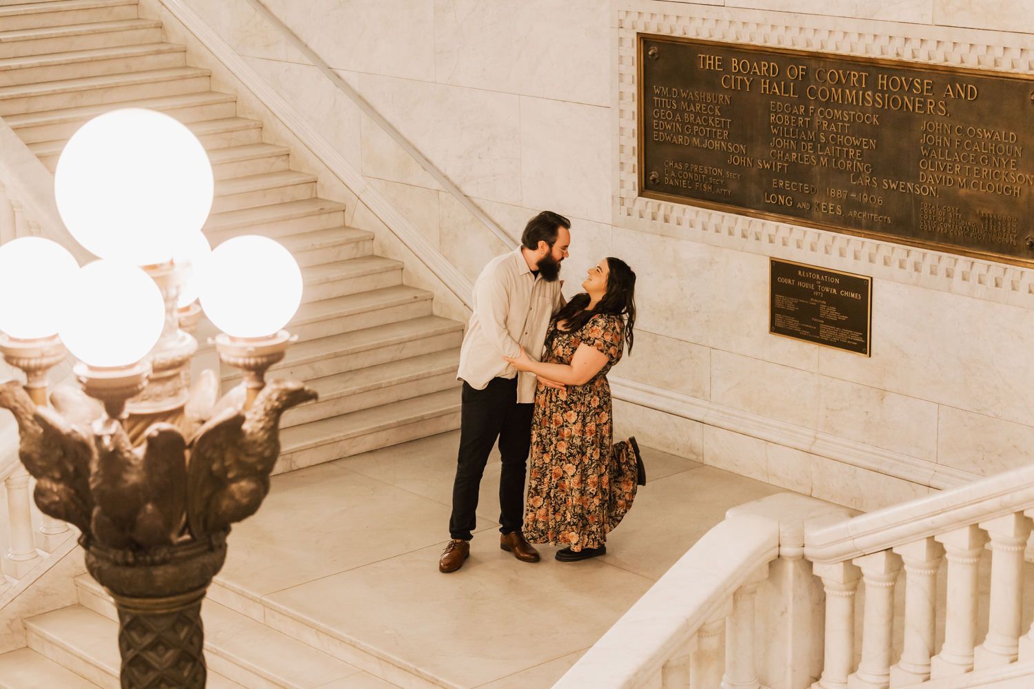 A couple stands intimately on an elegant staircase with ornate lighting and vintage architectural details in sepia tones.
