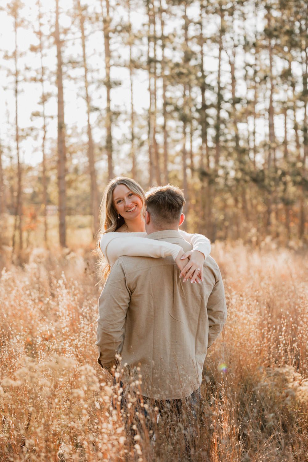 A romantic couple embraces in a sunlit field of tall grass with pine trees in the background.