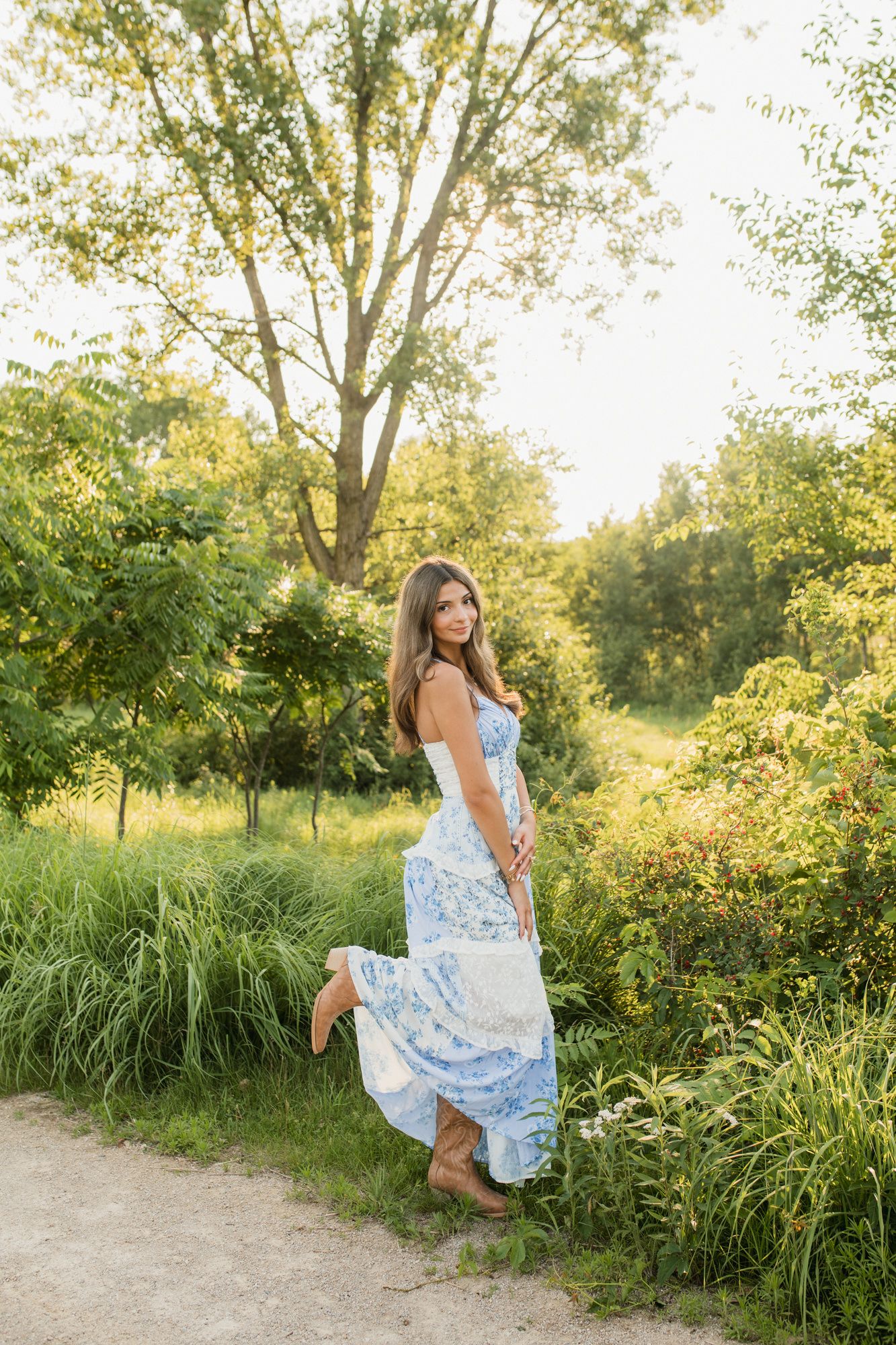 Woman in blue floral maxi dress posing on a dirt path surrounded by lush green trees and foliage in golden sunlight.