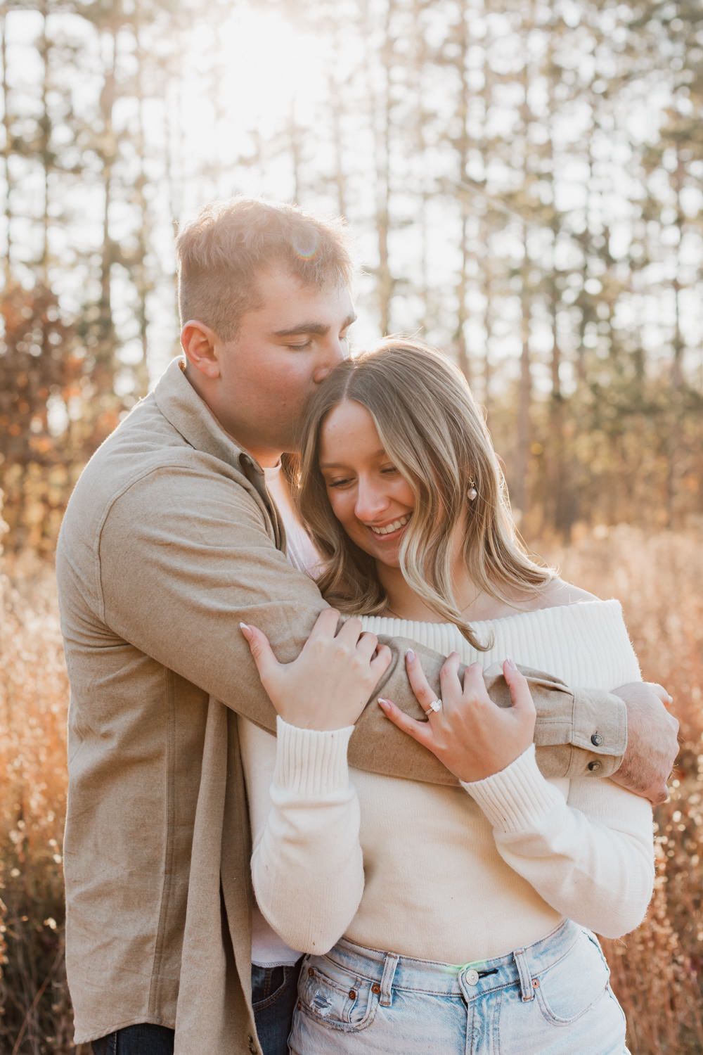A couple in casual fall attire sharing intimate moments in a field with golden grass and pine trees in the background.