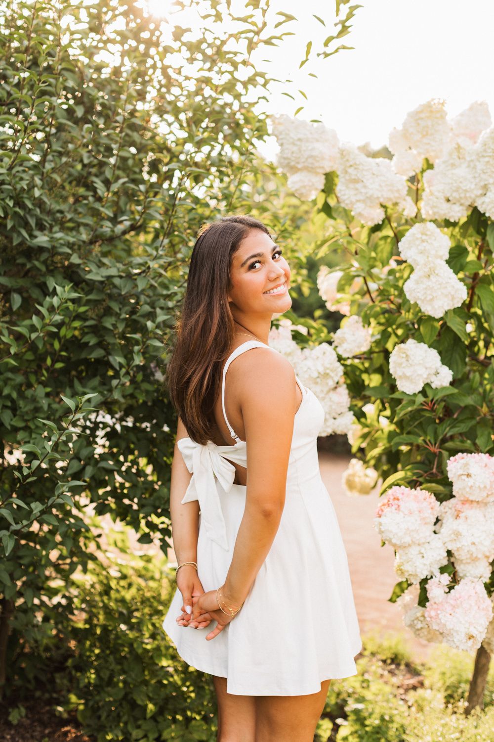 Golden hour portrait in a white dress with bow detail among flowering garden bushes.