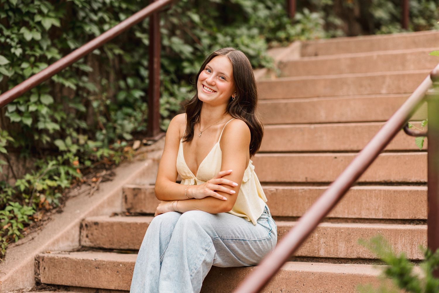 A person in casual summer attire sits and poses on outdoor wooden steps surrounded by lush greenery.