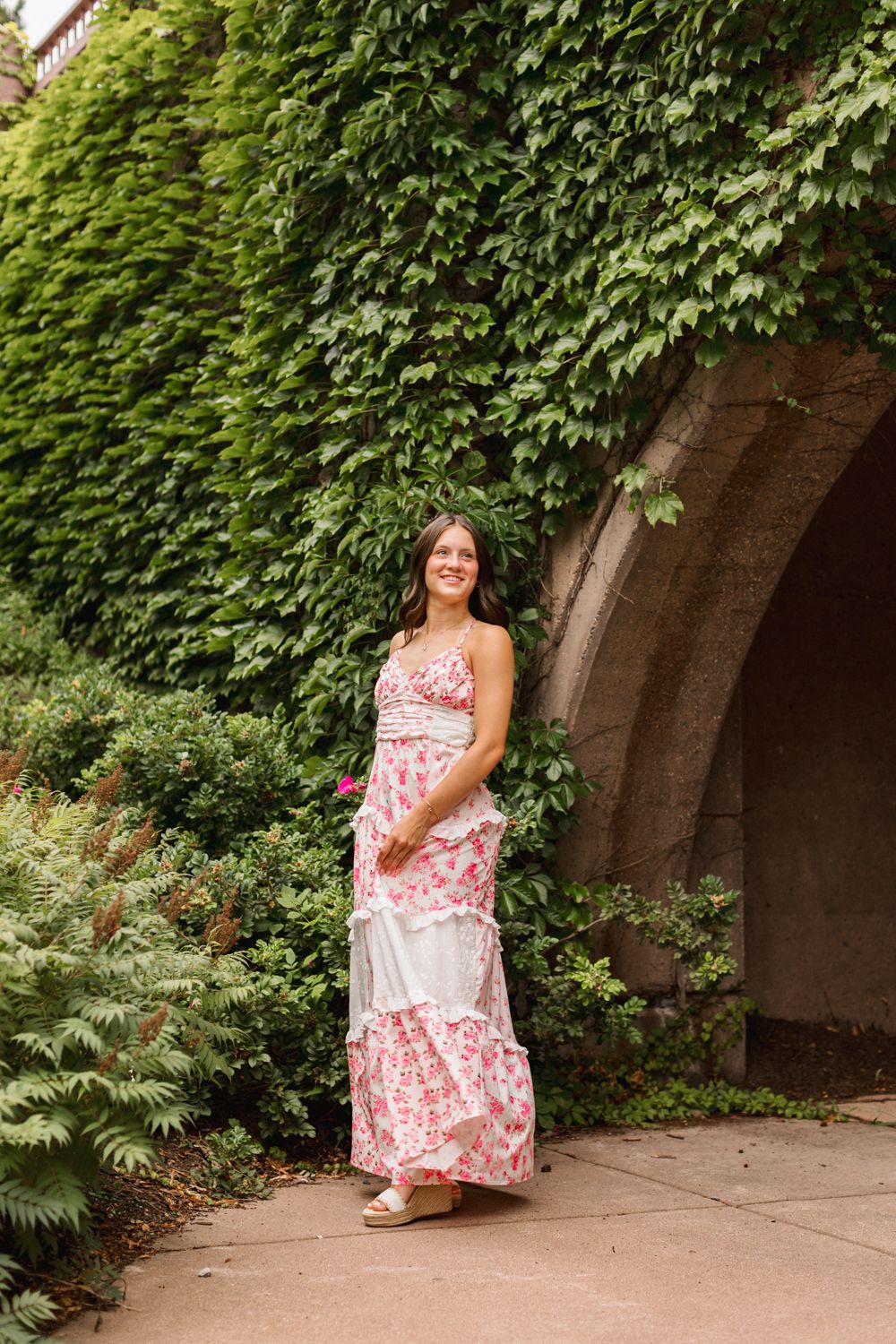 Young woman in a pink and white floral maxi dress poses by an ivy-covered archway in a garden setting.