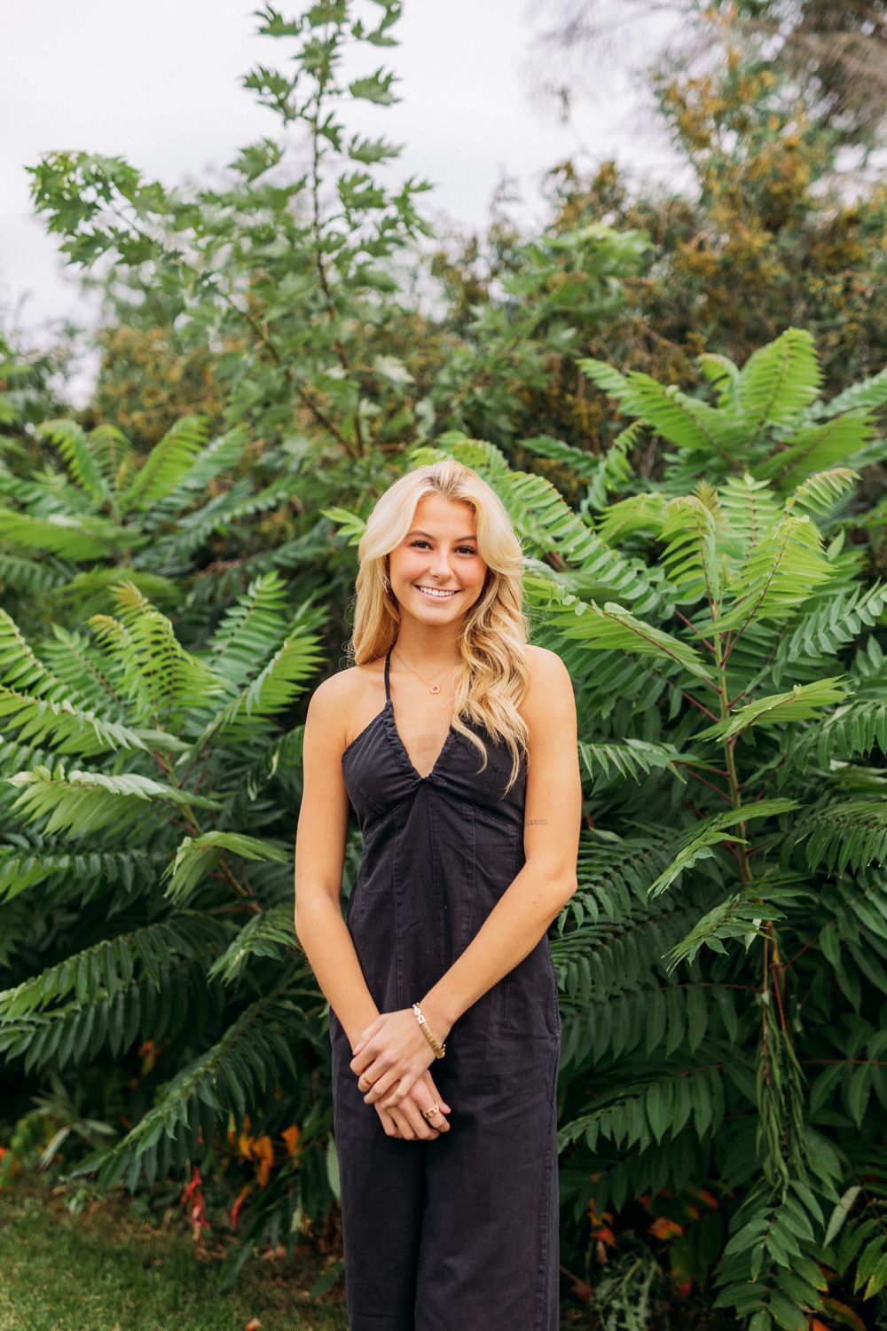 A figure in a black dress poses against lush green ferns in an outdoor garden setting.