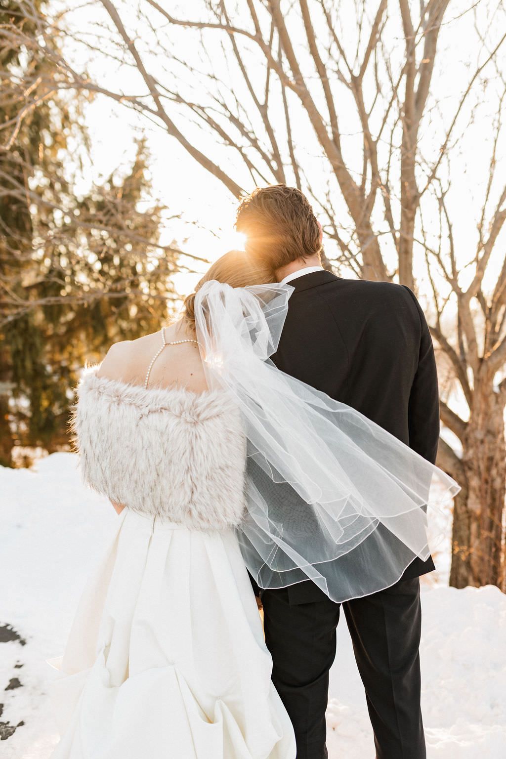 A bride's white dress flows elegantly against snowy winter scenery with bare trees.