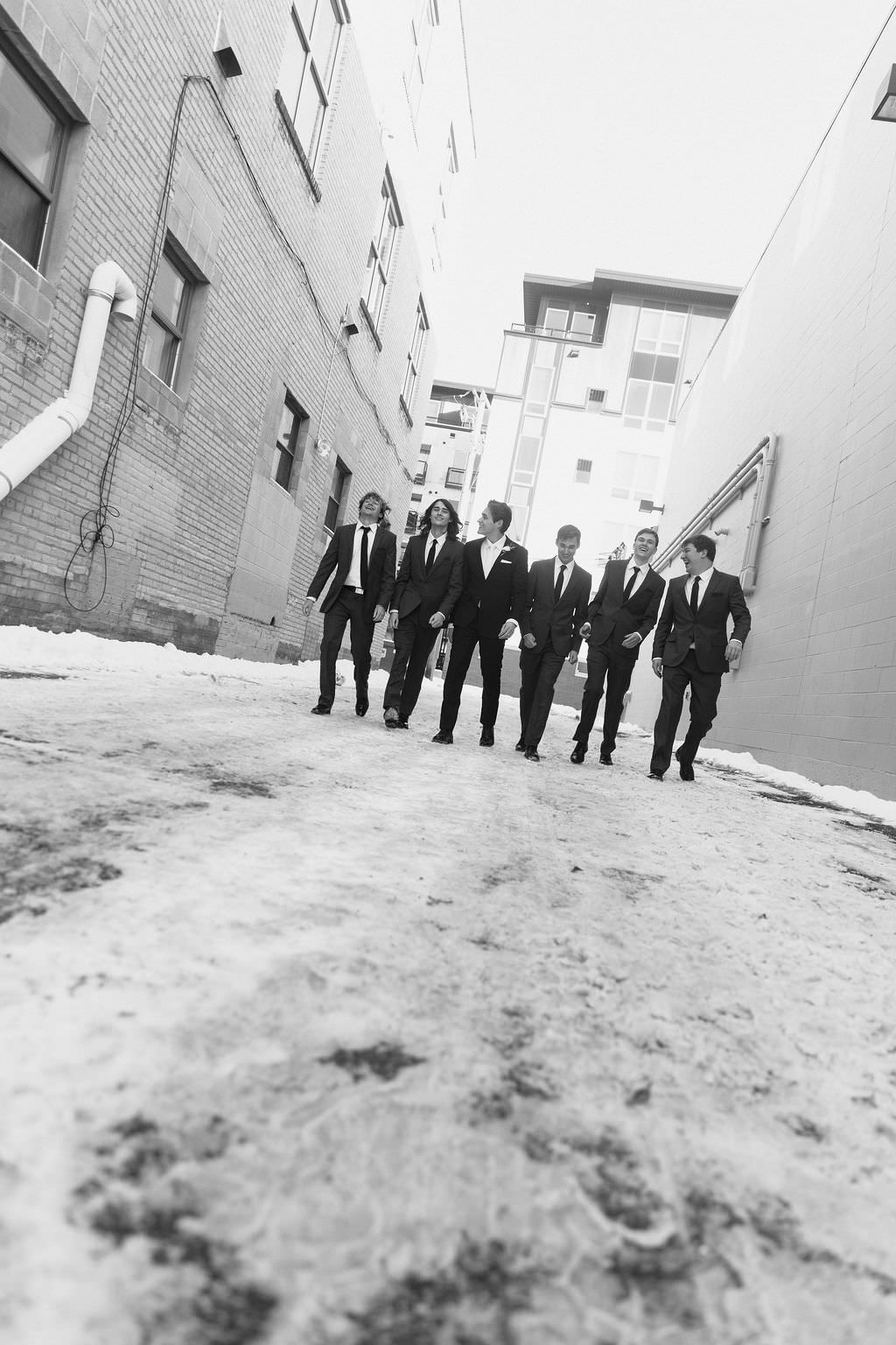 Wedding party in formal attire walks along brick wall in dramatic black and white photograph.