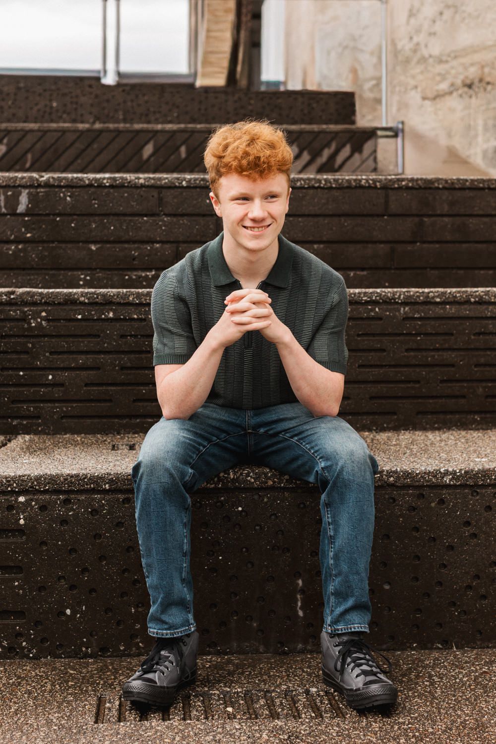 A figure in dark clothes sits on concrete steps against a black wooden wall with a contemplative pose.