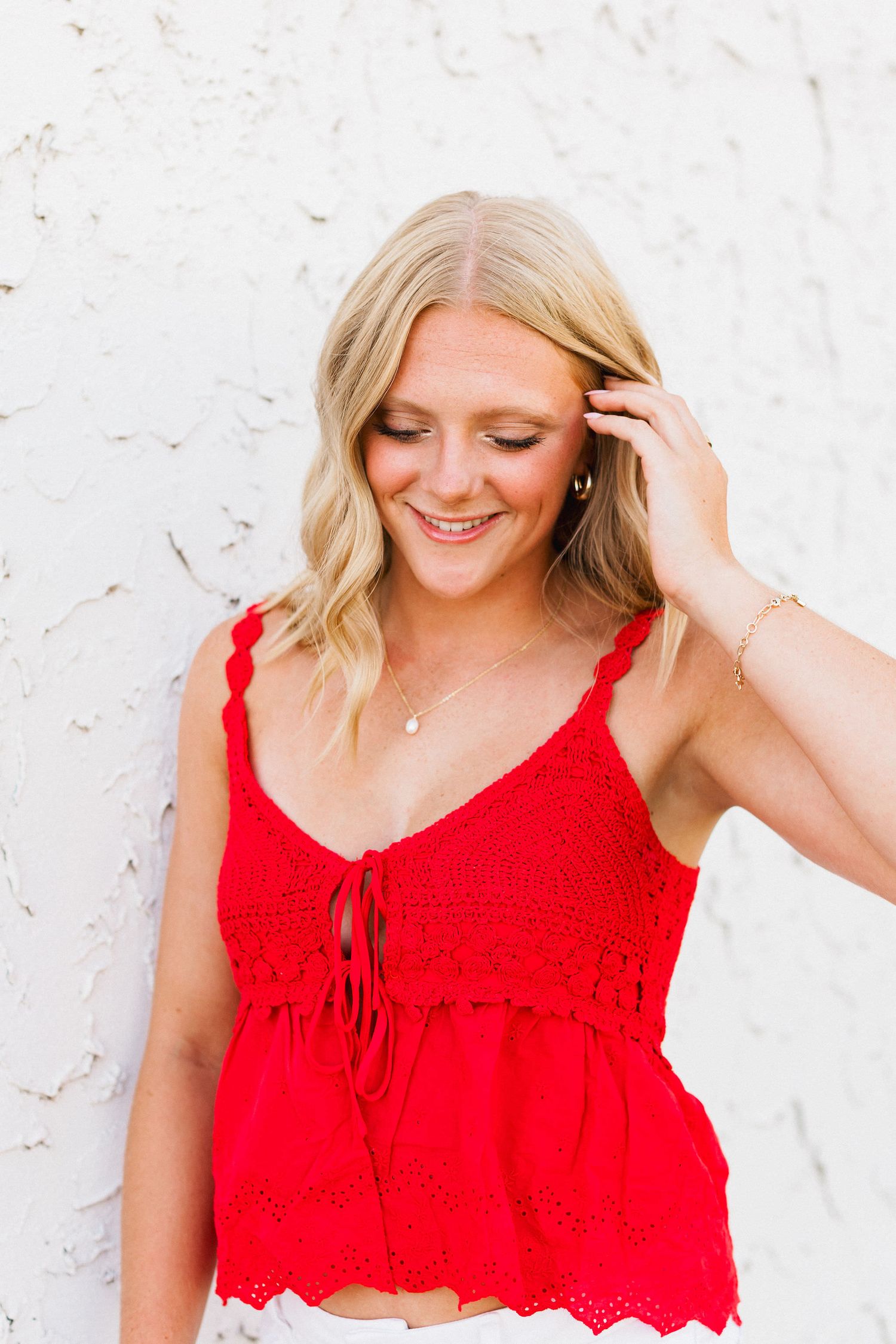 Woman in a red sleeveless top smiles while adjusting her hair against a white wall background.