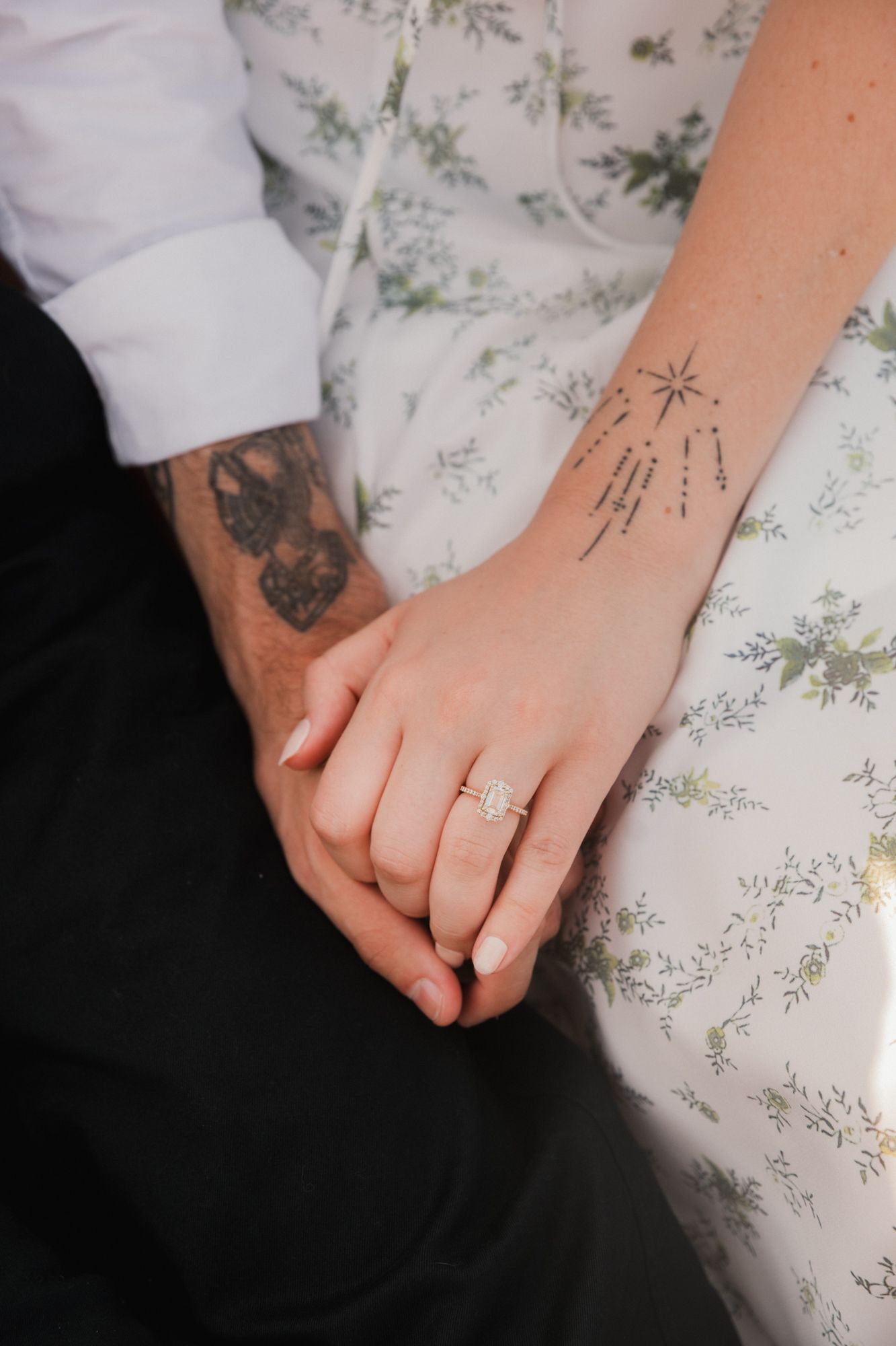 Close-up of couple holding hands, woman wearing diamond engagement ring, both with tattoos visible.