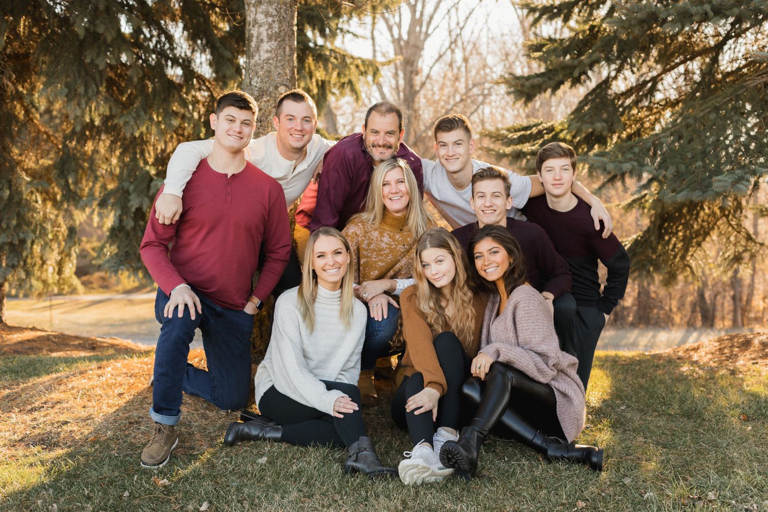 Large family group portrait outdoors among pine trees in autumn, smiling together on green grass.