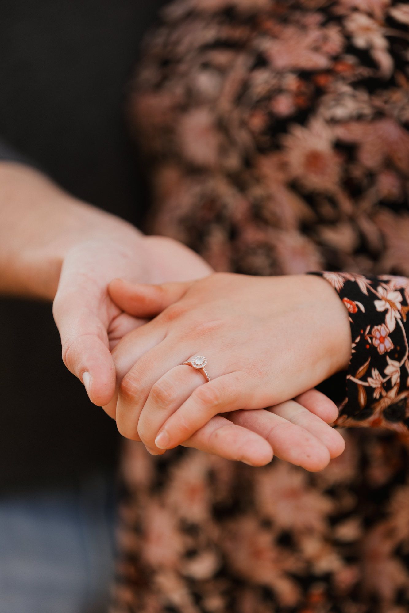 Close-up of couple holding hands displaying a delicate diamond engagement ring outdoors.