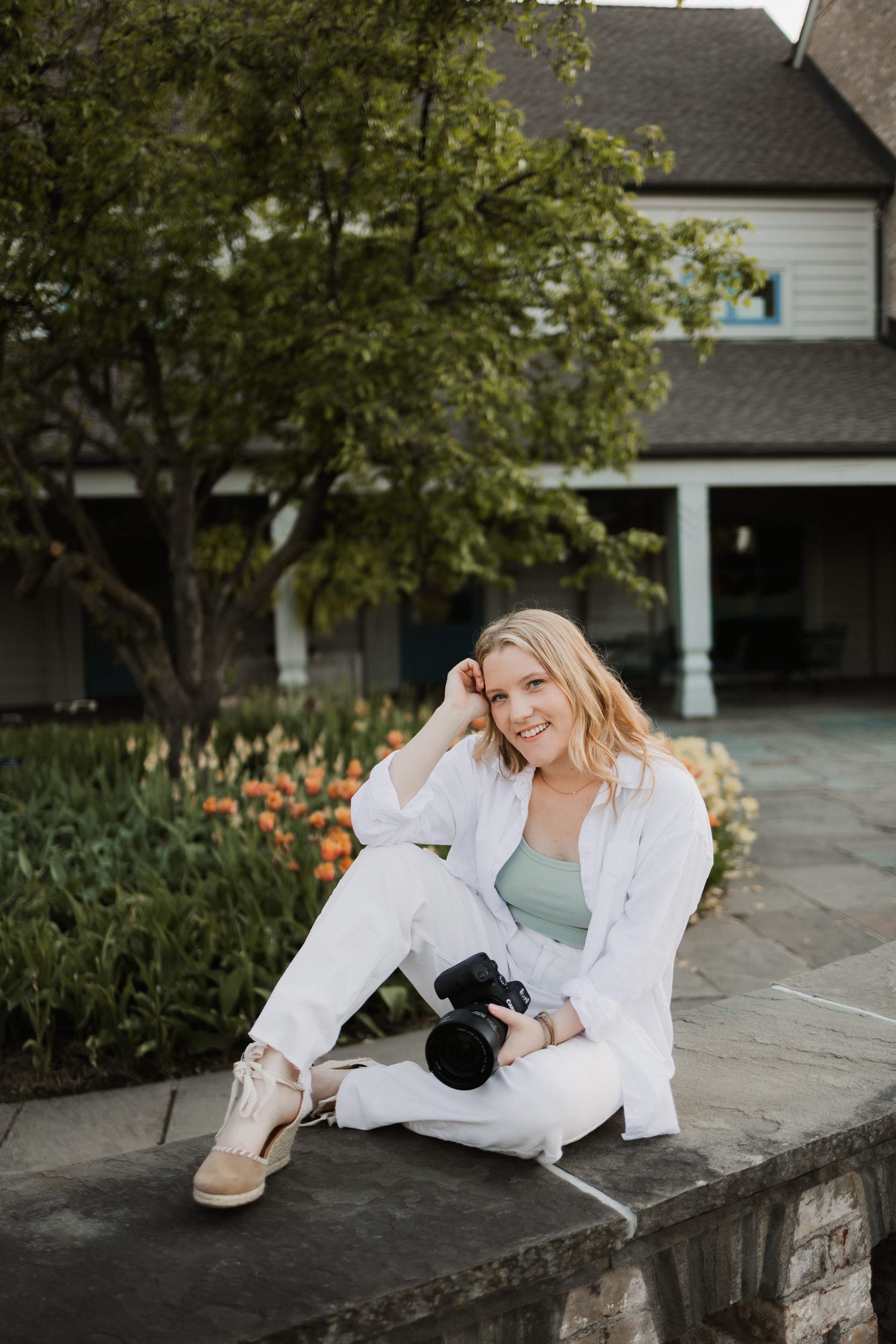 Smiling blonde woman in white outfit sitting outdoors holding a camera with colorful flowers and house in background.
