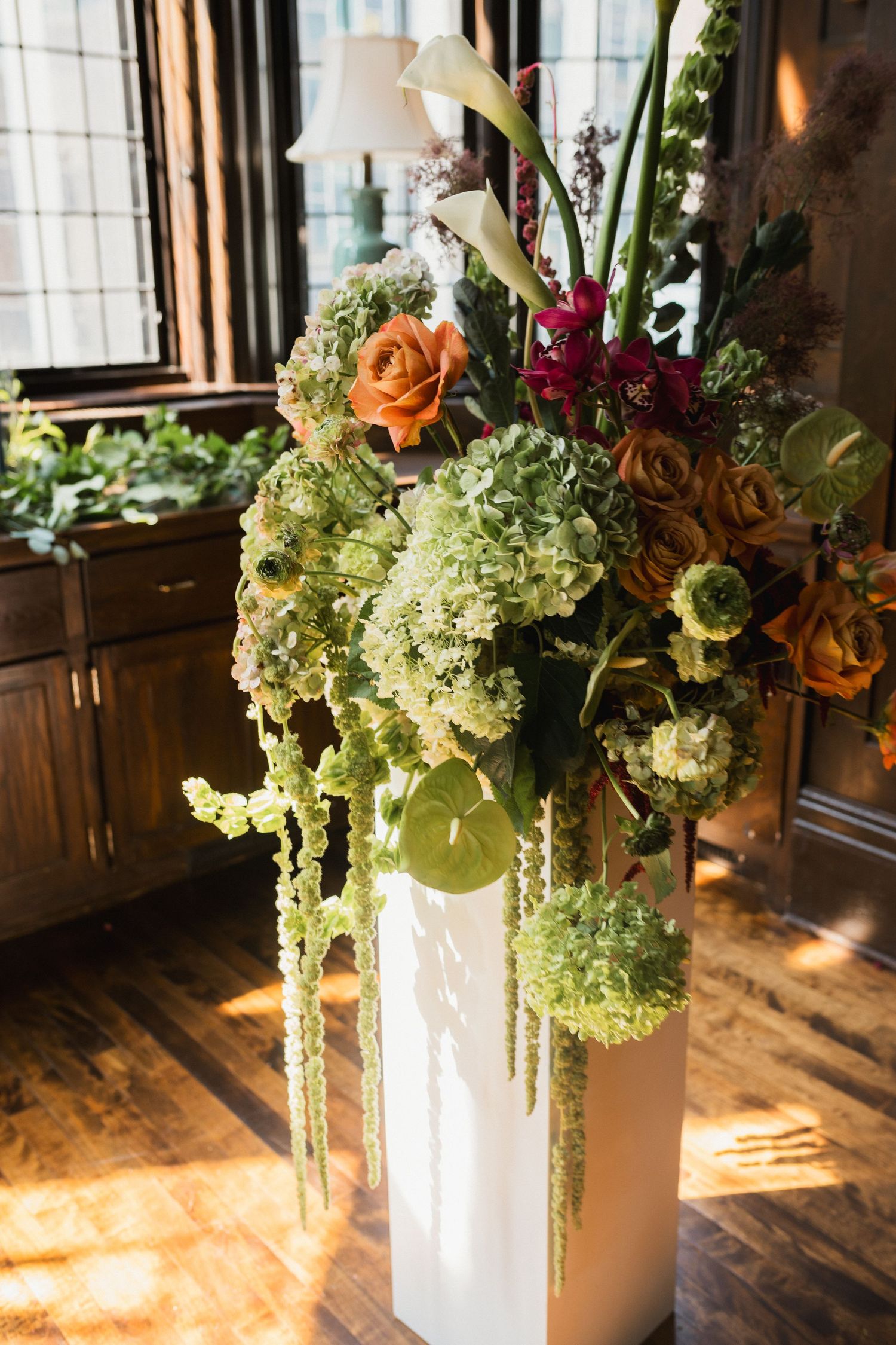 Elegant floral arrangement with green hydrangeas, orange roses, and white calla lilies on a white pedestal in a wood-paneled room.