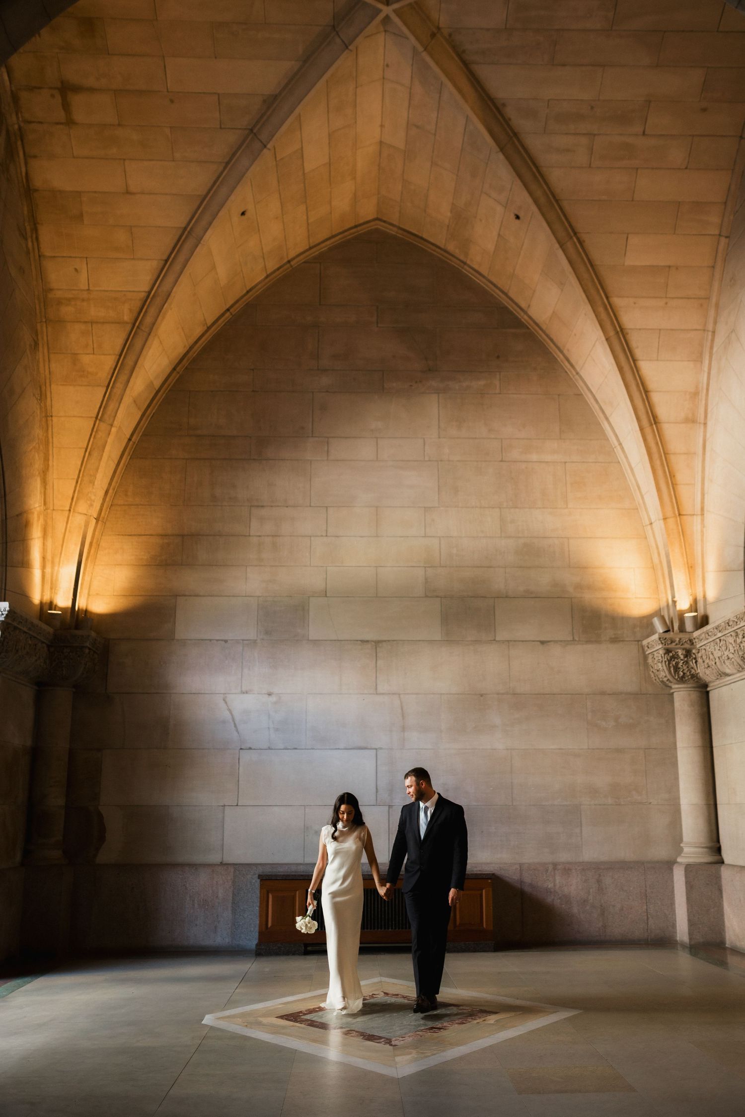 Bride and groom hold hands walking through grand Gothic stone archway in elegant wedding attire.