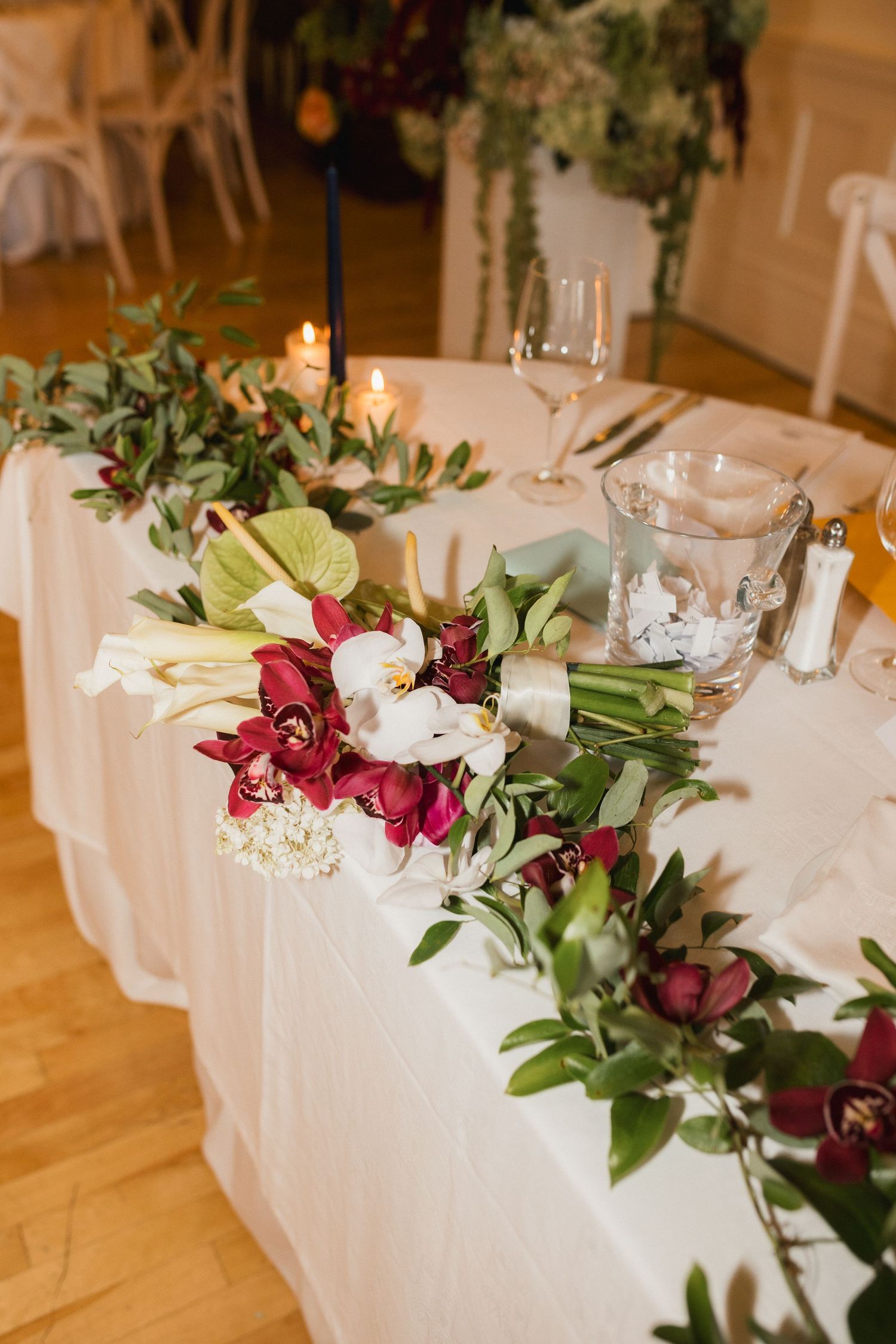 Wedding reception head table decorated with burgundy orchids, white flowers, and green garland centerpiece.