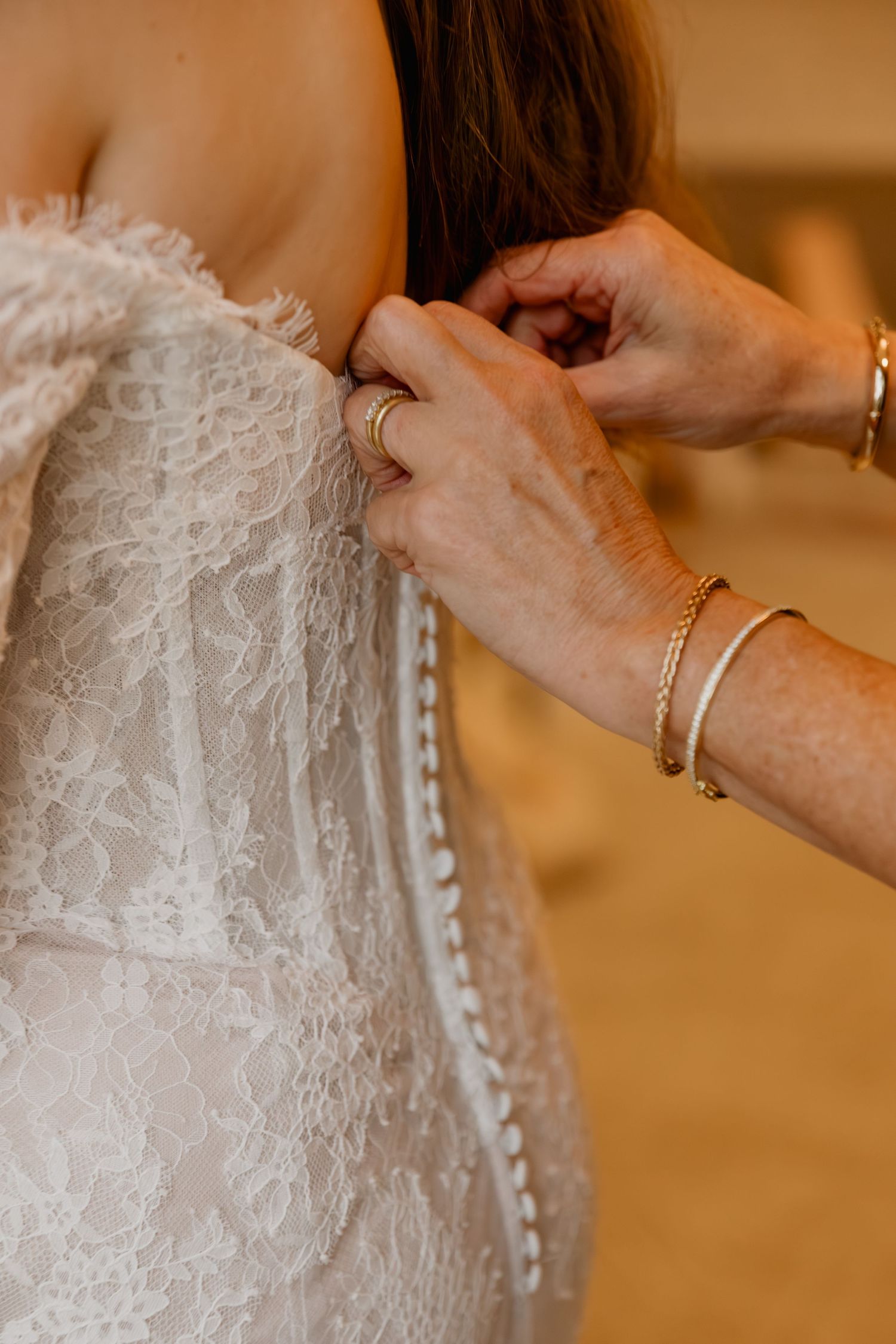 Close-up of a bride getting her lace wedding dress buttoned up by a woman wearing gold bracelets.