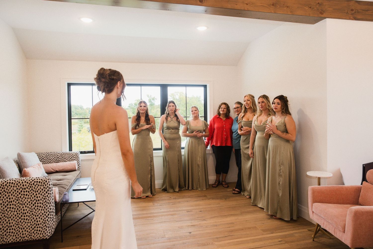 Bride in white gown reveals wedding dress to bridesmaids in sage green dresses during emotional first look.