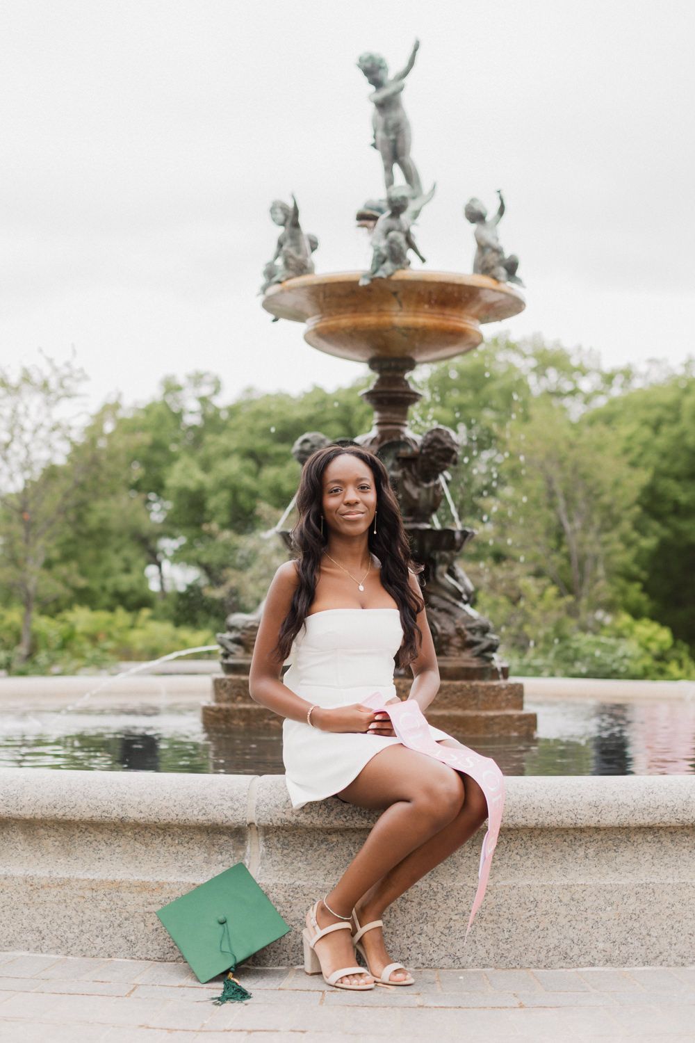 Graduate sits by ornate fountain in white dress with green graduation cap for celebratory photo session.