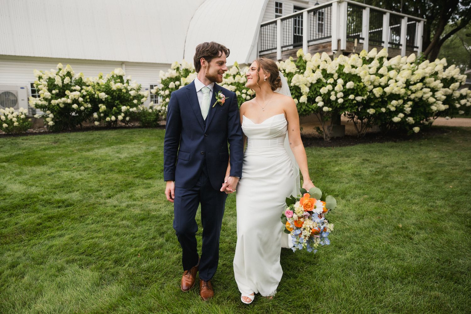 Bride and groom walking hand in hand on green lawn with white hydrangeas blooming behind them on their wedding day.