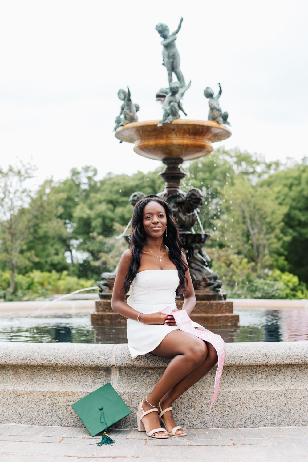 Graduate sitting by ornate fountain holding green graduation tassel and cap.