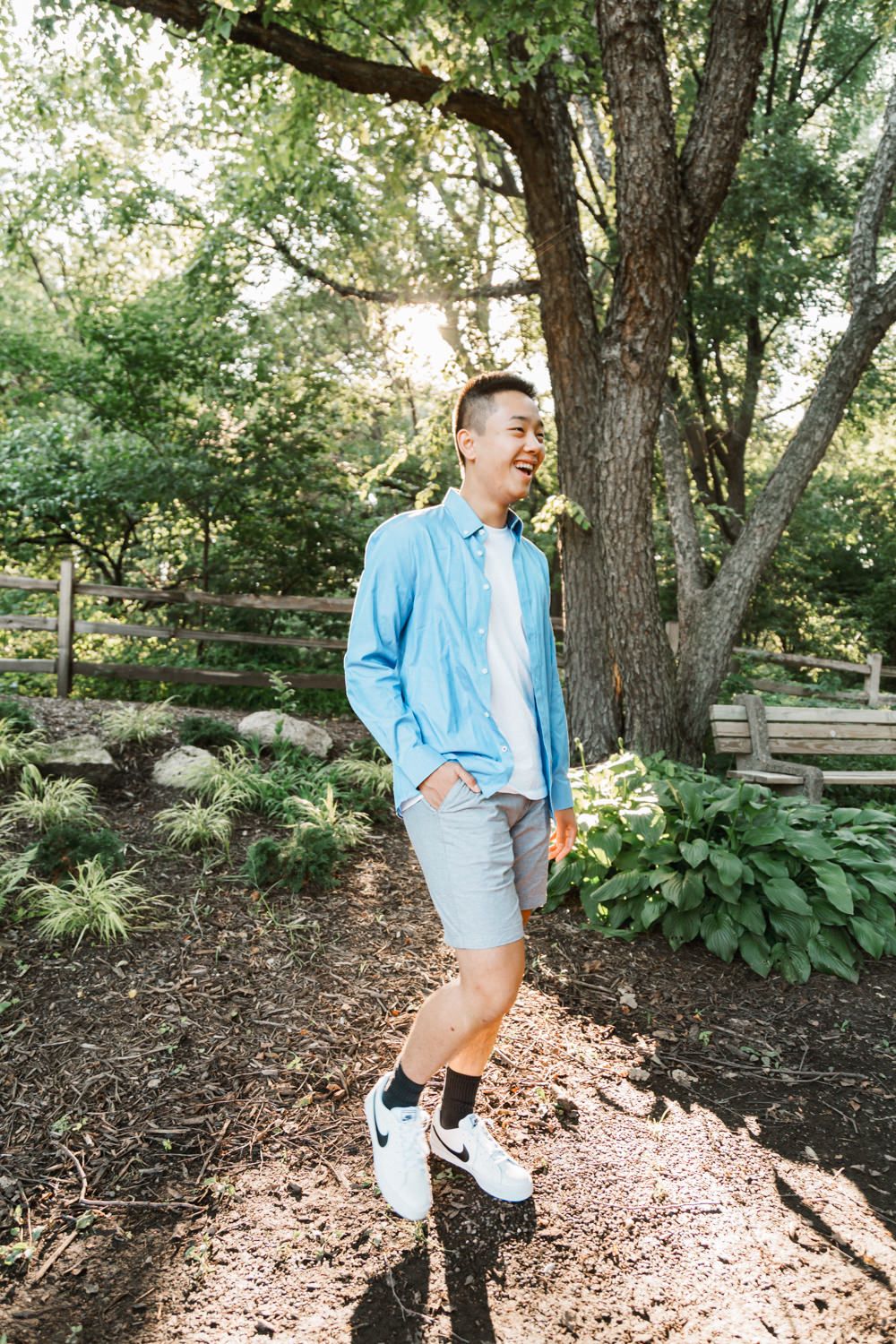 Someone in casual blue attire poses playfully near trees and ground foliage in a garden setting.