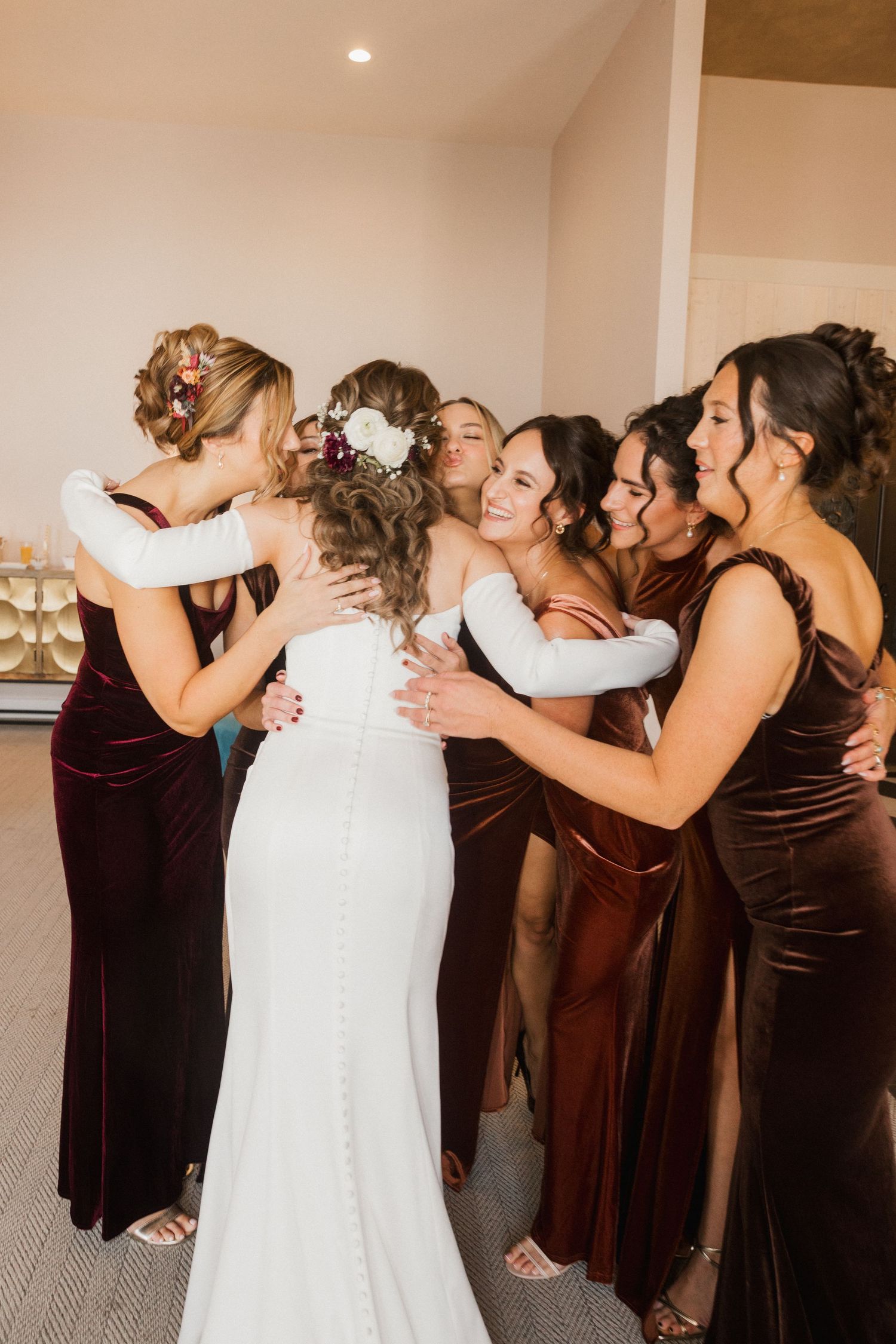 Bride in a white fitted gown hugs her bridesmaids in burgundy velvet dresses during a joyful group embrace before the wedding.