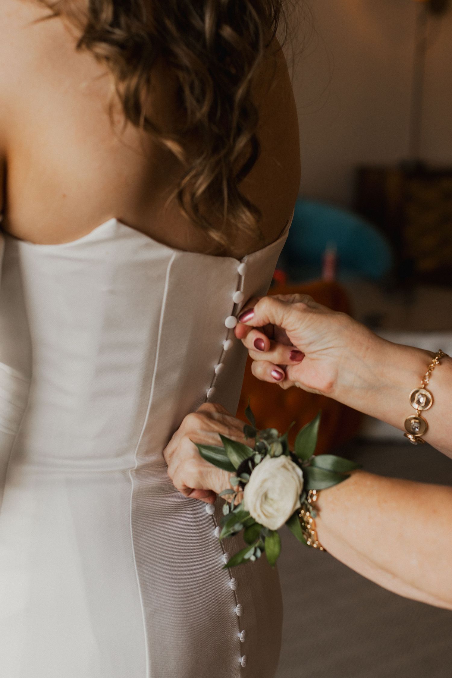 Hands fasten the buttons on the back of a white strapless wedding gown, with a white rose and greenery wrist corsage.