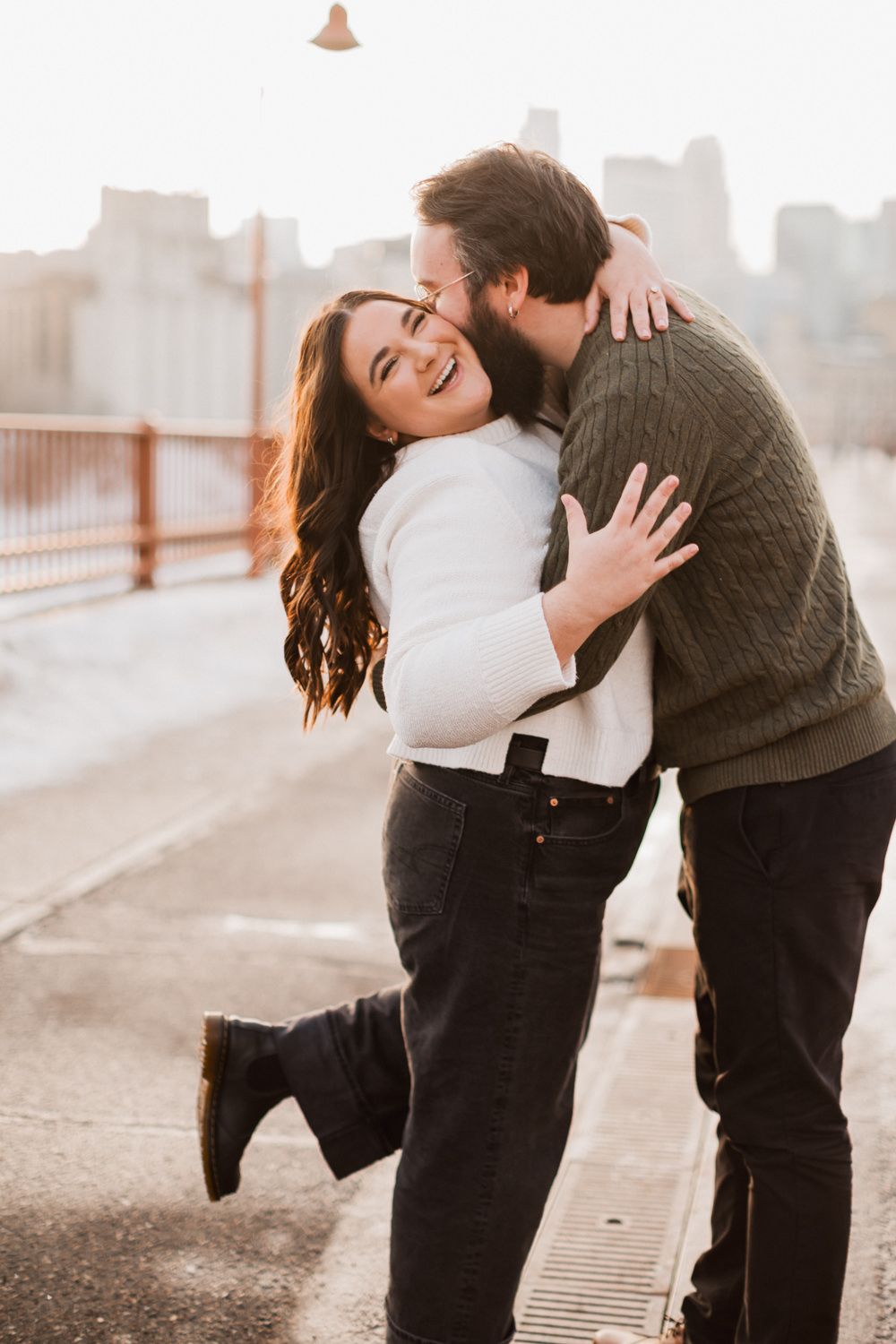 Joyful couple celebrating their engagement on a bridge with urban backdrop.