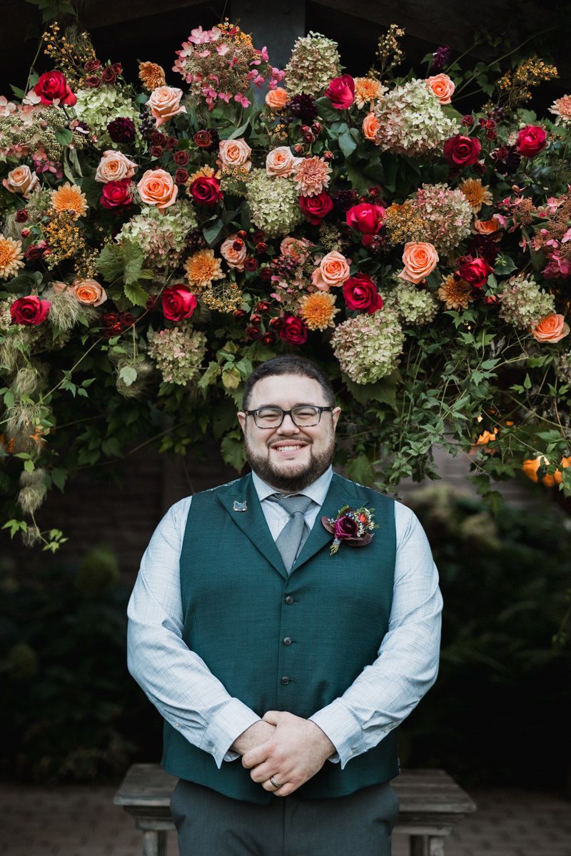 Person in green vest smiles under stunning floral arch with roses and hydrangeas.