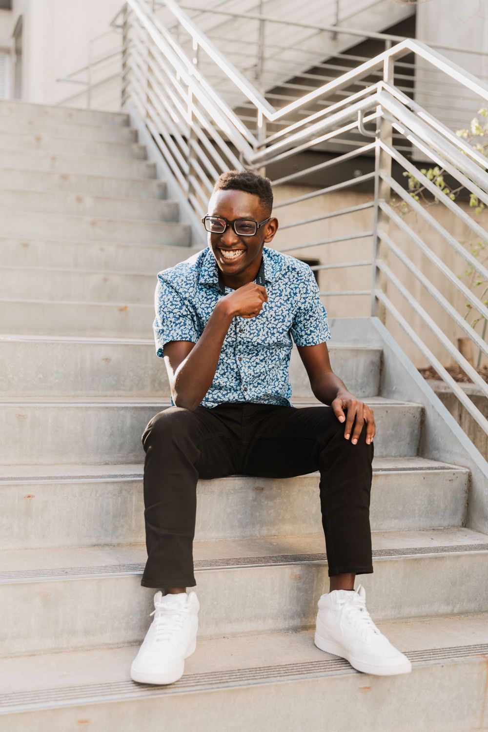 A person in a patterned blue shirt and black pants sits casually on concrete stairs while smiling.