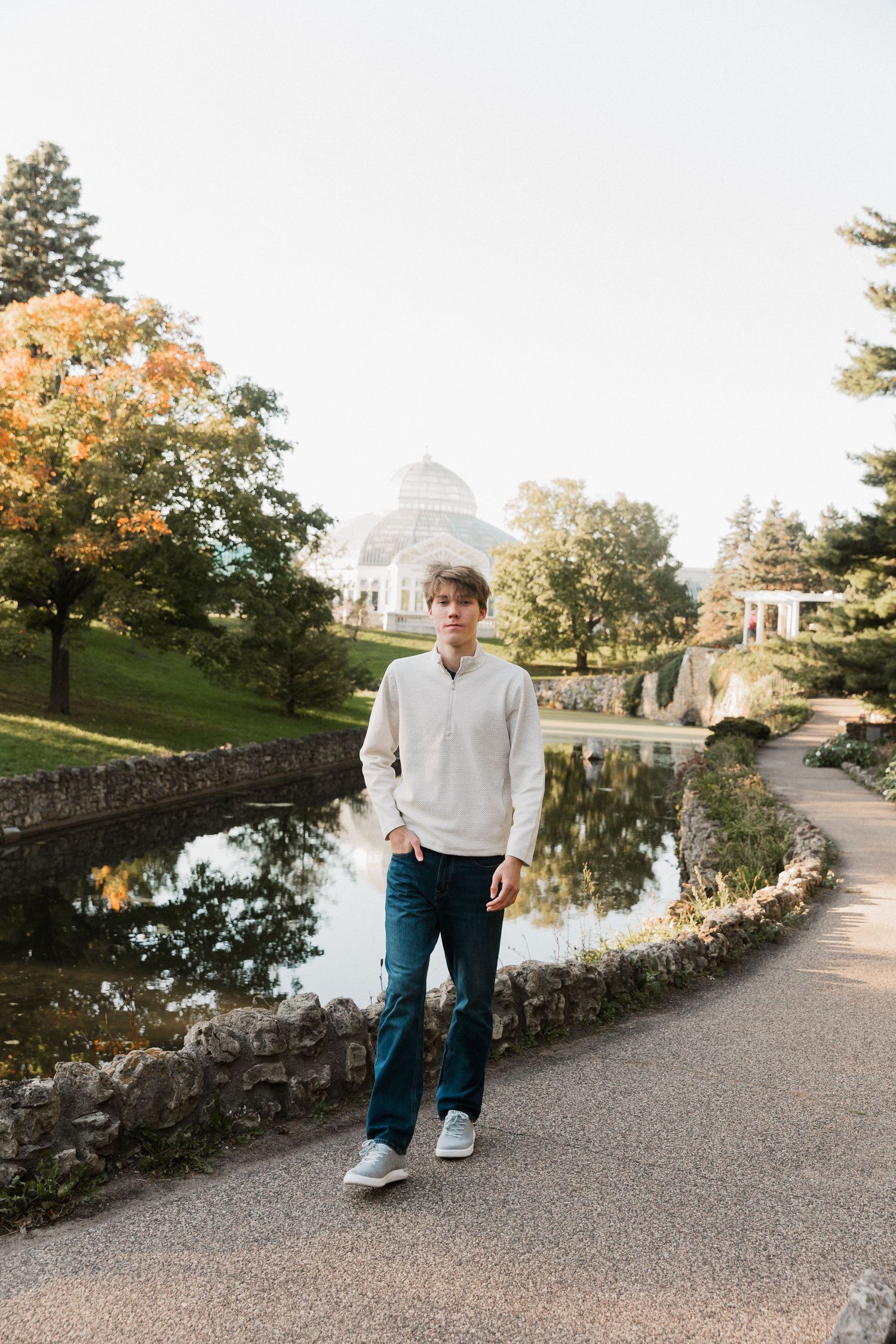 A scenic autumn path along a stone wall and reflecting pond leads to a white gazebo in a tranquil garden setting.