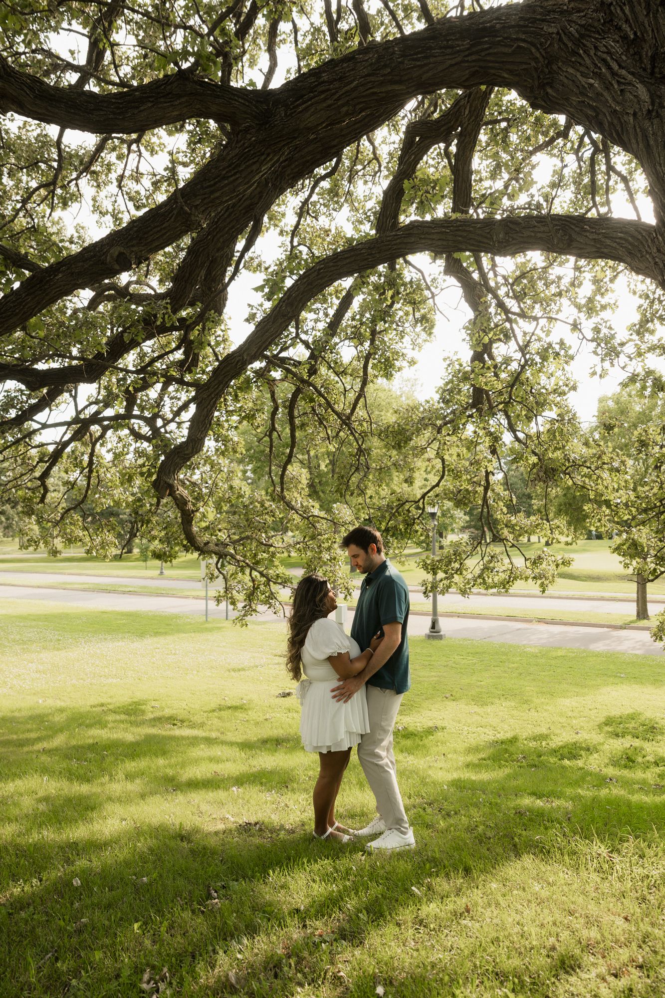 Couple embracing under a large oak tree in a lush green park, woman in white dress, man in green shirt.
