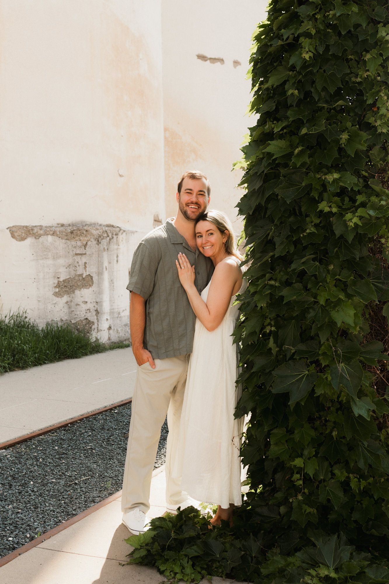 A couple embraces near an ivy-covered pillar beside a rustic white wall in warm sunlight.