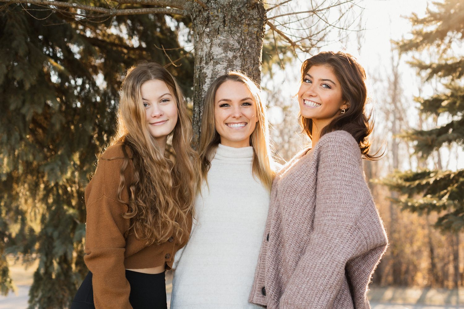 Three smiling young women pose outdoors near evergreen trees in cozy sweaters on a sunny winter day.
