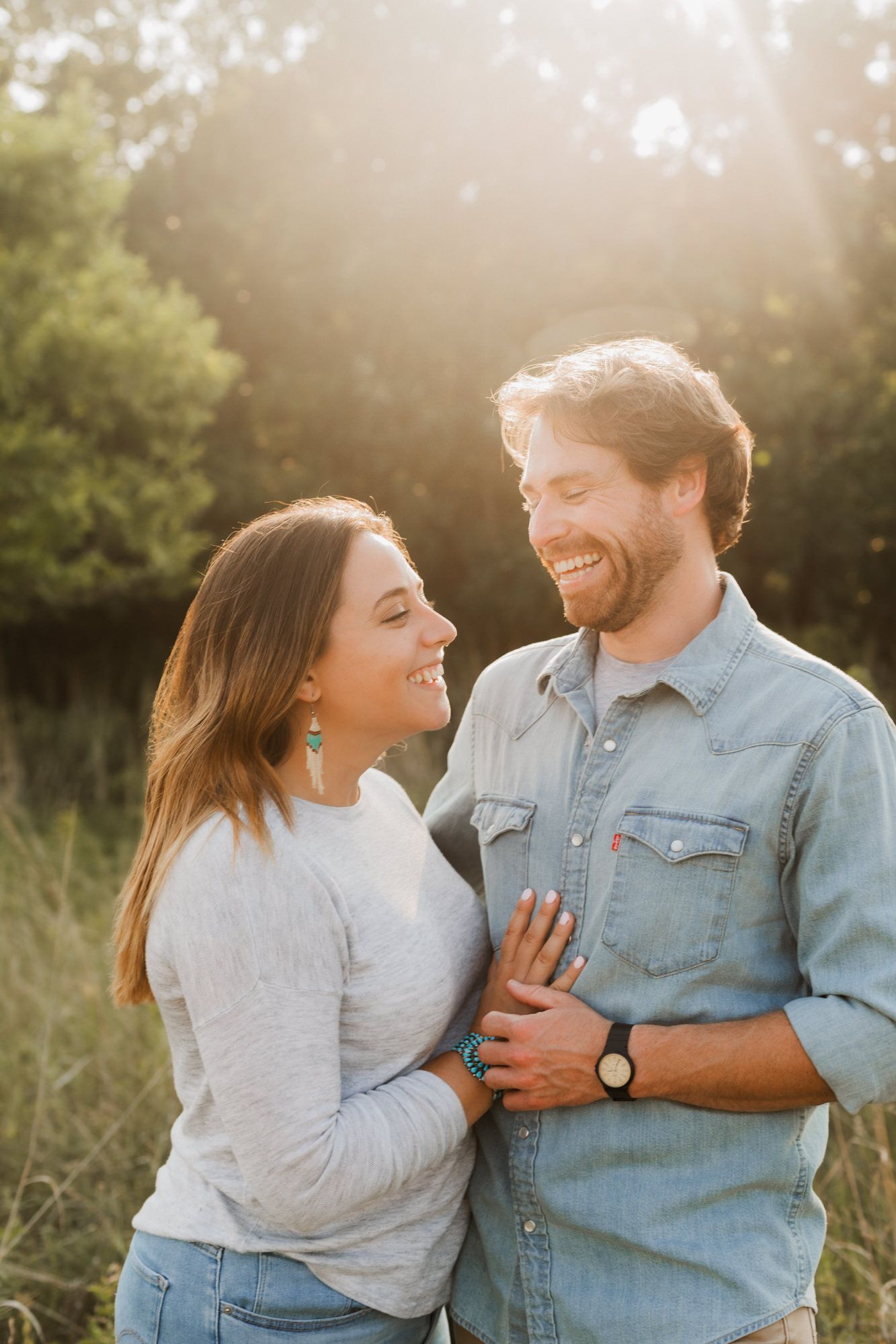 Smiling couple embracing in a sunlit field, woman in gray sweater with green earrings, man in denim shirt wearing a watch.