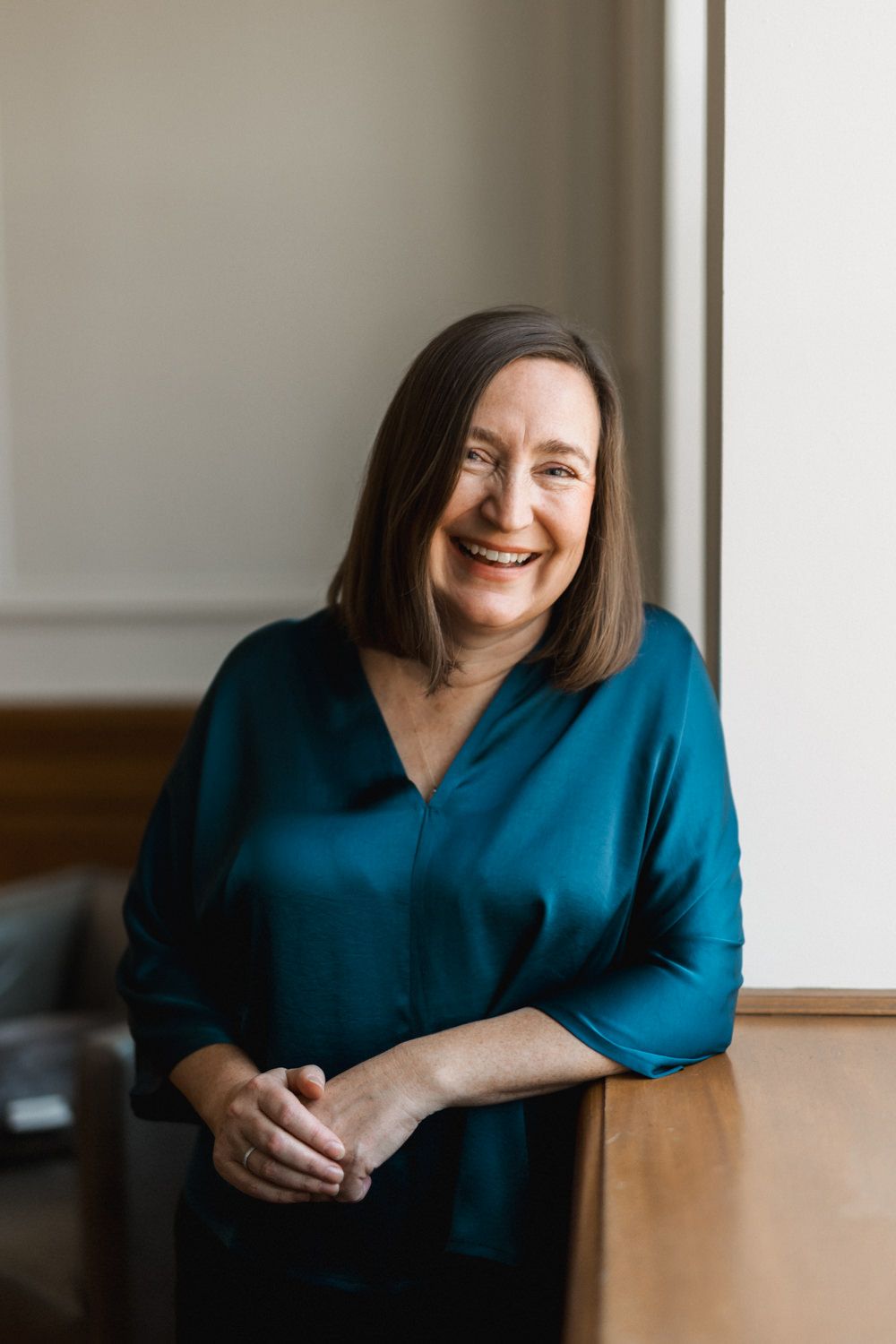 Smiling middle-aged woman with brown hair wearing a teal blouse, leaning against a wooden table in a bright office.