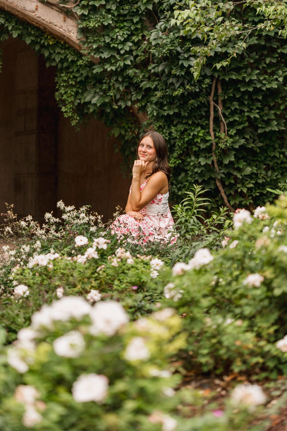 A person sits thoughtfully among blooming white roses and greenery near an ivy-covered wall.